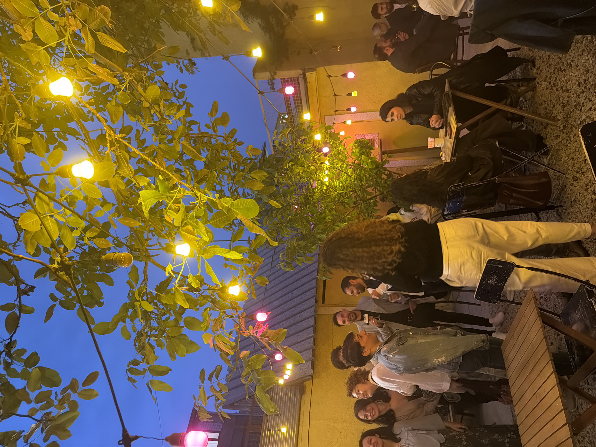 The image depicts an outdoor dining area, likely in Yerevan, Armenia, given the presence of Armenian-speaking individuals and the architectural style. A group of people is gathered at tables, some seated and some standing. There's an arrangement of trees and foliage interspersed with string lights, creating a warm, convivial atmosphere. The lights are a mix of yellow and red hues. Some people are engaged in conversation. One person is sitting alone at a table. The ground is covered with gravel. The time of day appears to be evening or late afternoon, with a visible darkening blue sky.