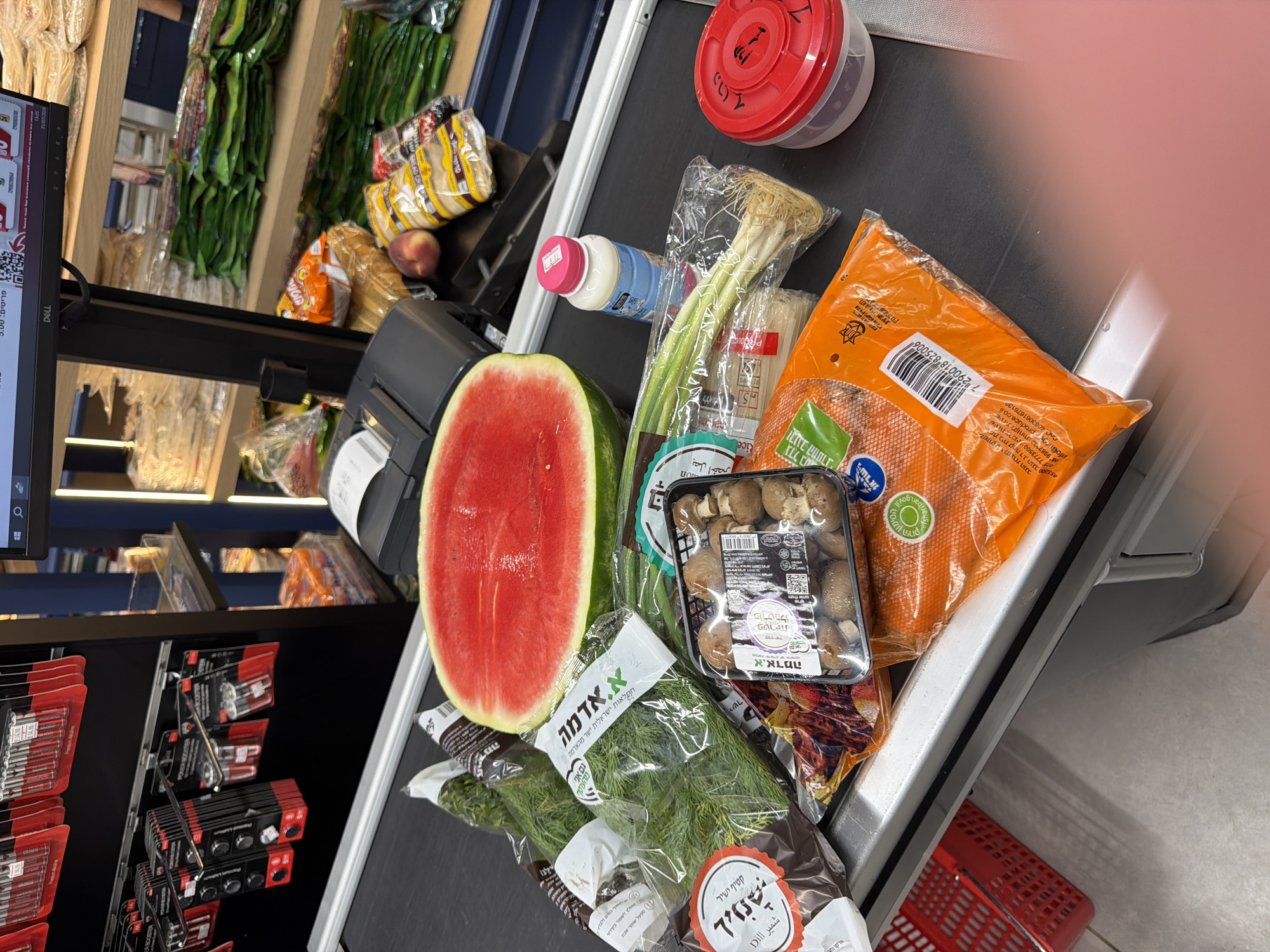 The image shows a conveyor belt at a supermarket checkout in Even Yehuda, Israel. A partially sliced watermelon is on the belt, alongside a container of mushrooms, a bunch of green onions, and several packages of herbs and other produce. A pink plastic container with a red lid is also visible. In the background, shelves are stocked with packaged goods, and a computer monitor displays a checkout interface. The overall scene depicts a typical grocery shopping experience at a checkout counter.