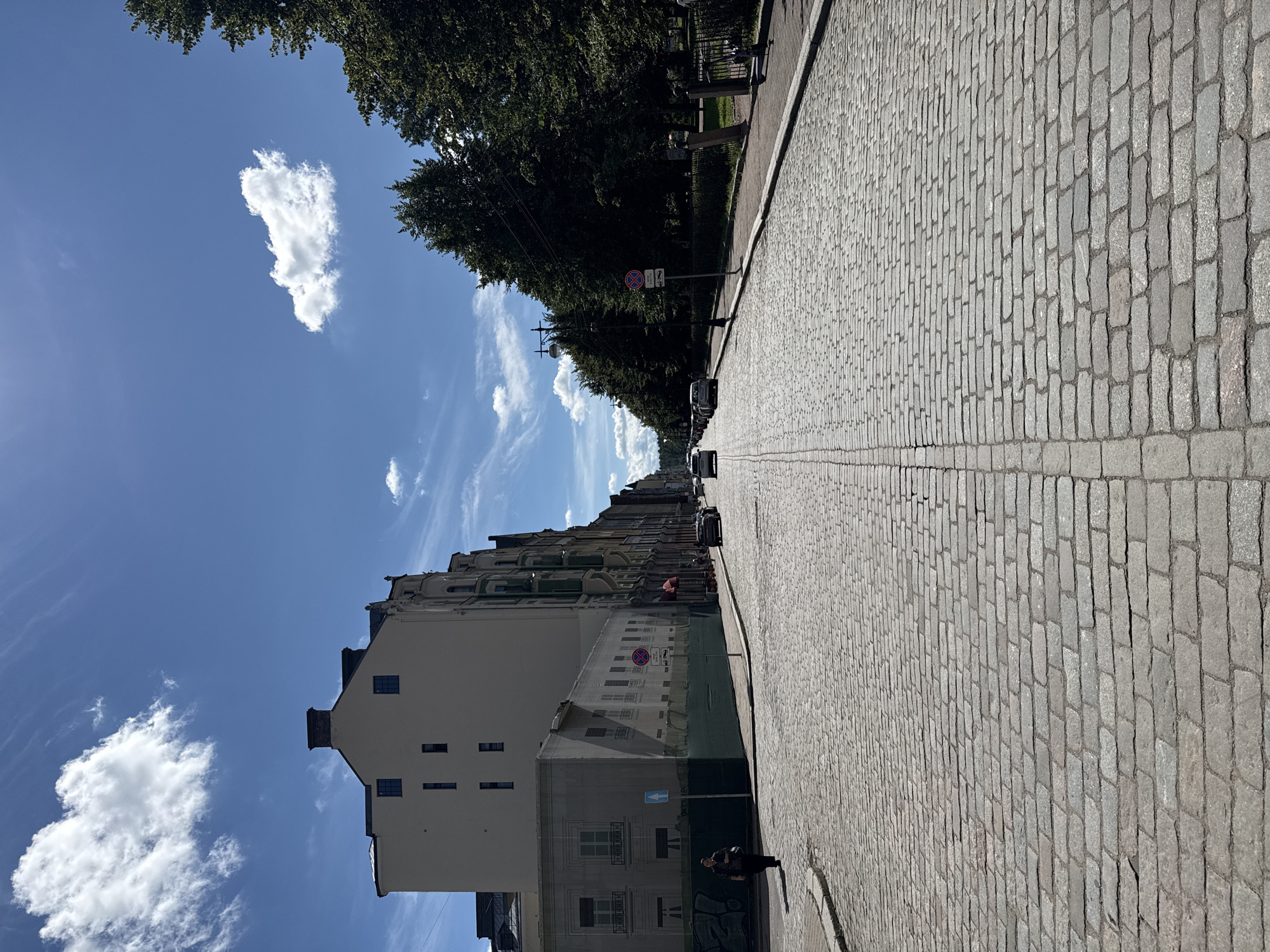 A straight, cobblestone street stretches into the distance under a bright blue sky with scattered white clouds. On the left, a tall, light-colored building with multiple windows dominates the foreground, partially obscured by a dark green tarp covering a construction area. A woman in dark clothing walks away from the camera on the left sidewalk. Further down the street, several cars are visible. On the right side of the street, a line of mature, leafy green trees forms a dense canopy, with a sidewalk running alongside it. A black metal fence separates the sidewalk from a park area. A black electric scooter is parked on the right sidewalk. Signage, including a red and white no stopping sign with a tow-away symbol, is visible along the street. The architecture suggests a European city, likely Vyborg, Russia, given its historical cobblestone streets and building styles.
