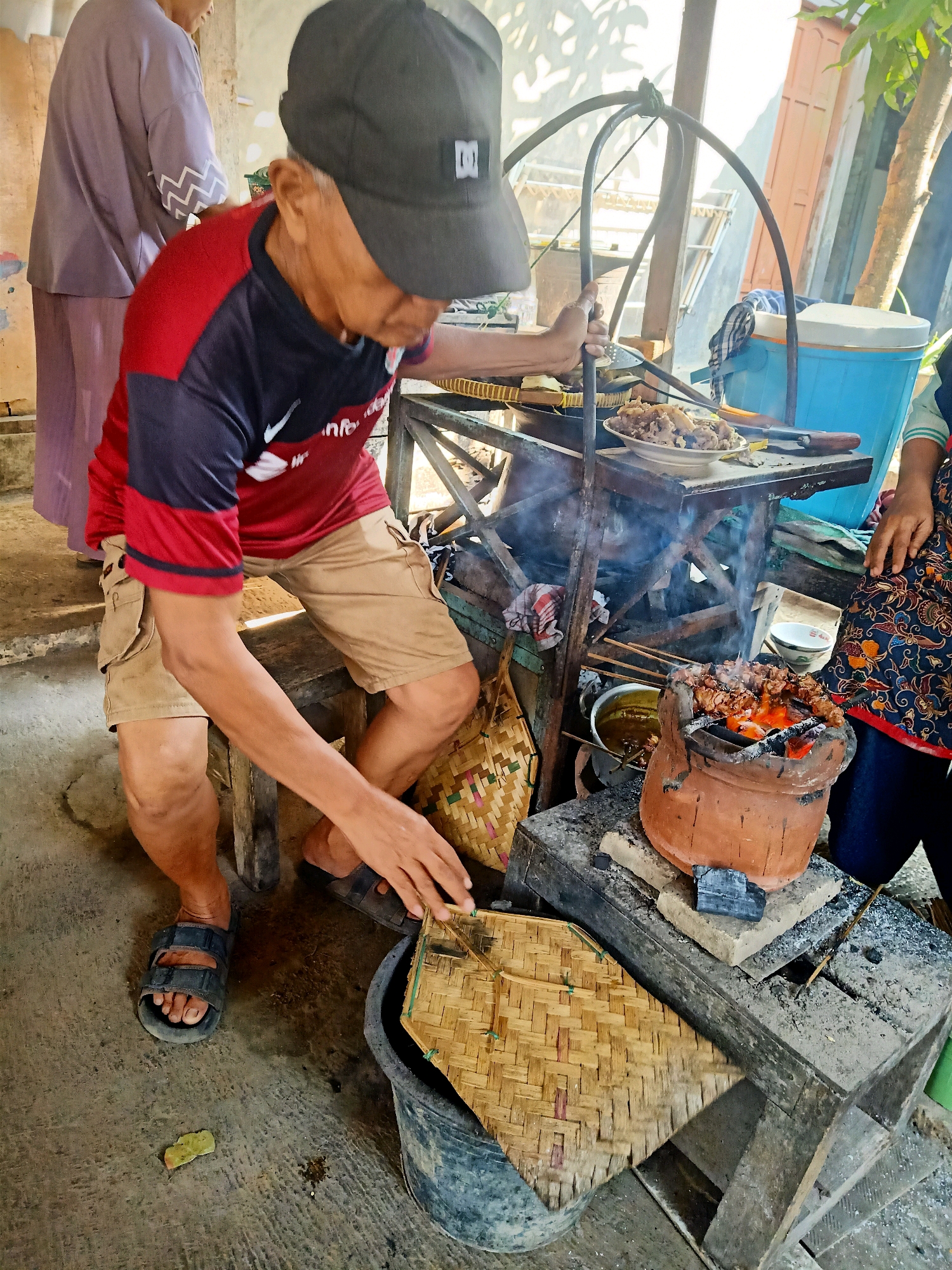 In Jatinom, Indonesia, an elderly man wearing a red and black shirt, khaki shorts, and a black baseball cap is seen grilling satay over a charcoal brazier. He is leaning over the grill, using a spatula to adjust the skewers. To his left, another person in a purple garment stands by. In the background, a blue cooler is visible. The scene appears to be an outdoor food stall.