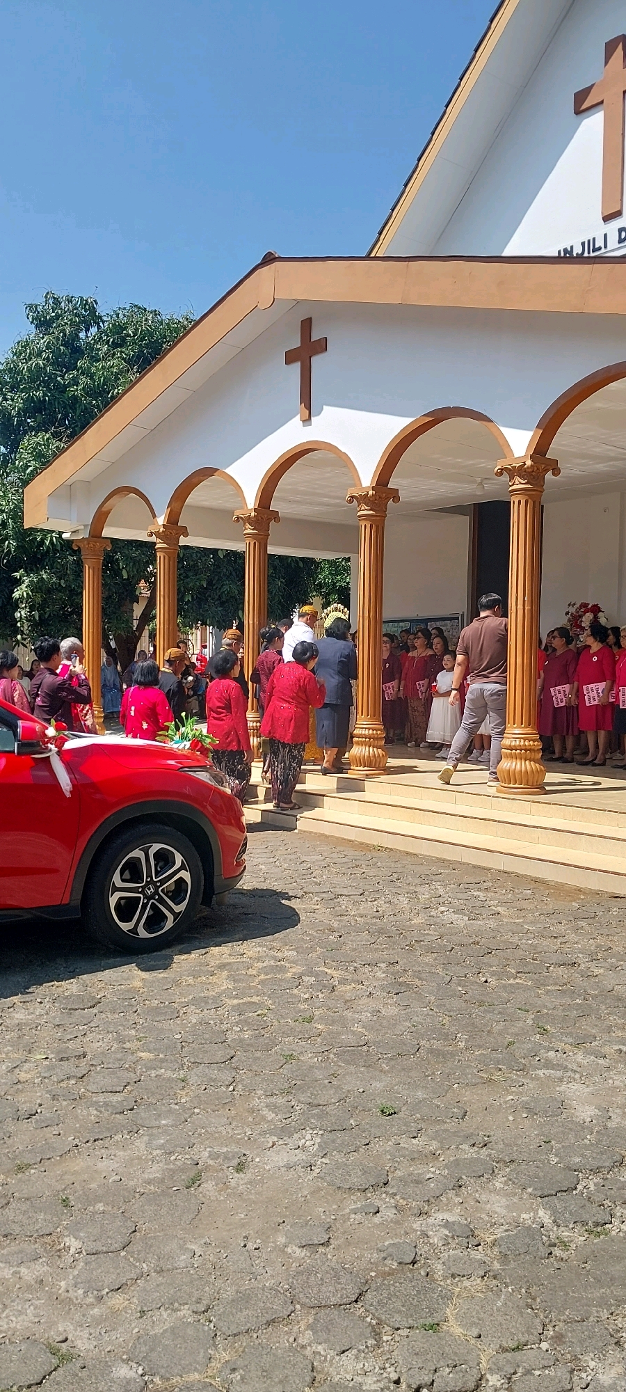 A group of people are gathered outside a white building with a brown-trimmed roof and arched entrances supported by decorative columns, identified as the GBI Jemaat Immanuel church in Keling, Indonesia. The scene appears to be a ceremony or event, with many individuals dressed in traditional clothing. A red car, adorned with a white ribbon, is parked to the left of the frame, facing away from the building. The ground is paved with hexagonal stones. The sky is clear and blue.