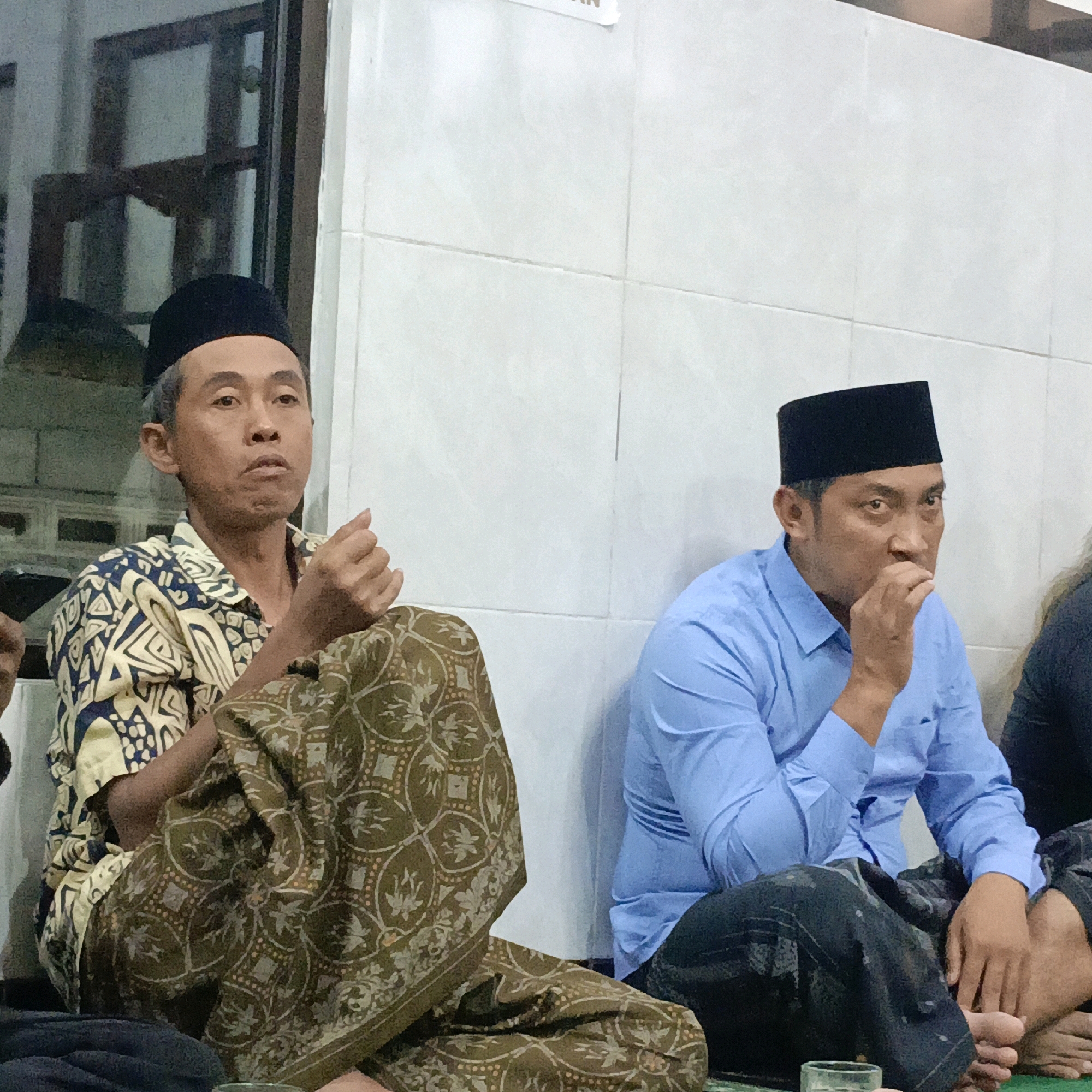 Two men in prayer caps and traditional clothing are seated on the floor indoors. The man on the left wears a black prayer cap and a patterned shirt with blue and yellow motifs, and a brown batik sarong draped around his legs. He is looking to his left with his hands resting on his lap. The man on the right wears a black prayer cap and a light blue long-sleeved shirt. He is looking forward and has his hand near his mouth. He is wearing dark pants, and his legs are crossed. In the foreground, a glass of clear liquid is visible on a green mat. The wall behind them is tiled with white square tiles. The scene appears to be in a residential or community setting. This image was captured in Salatiga, Indonesia.