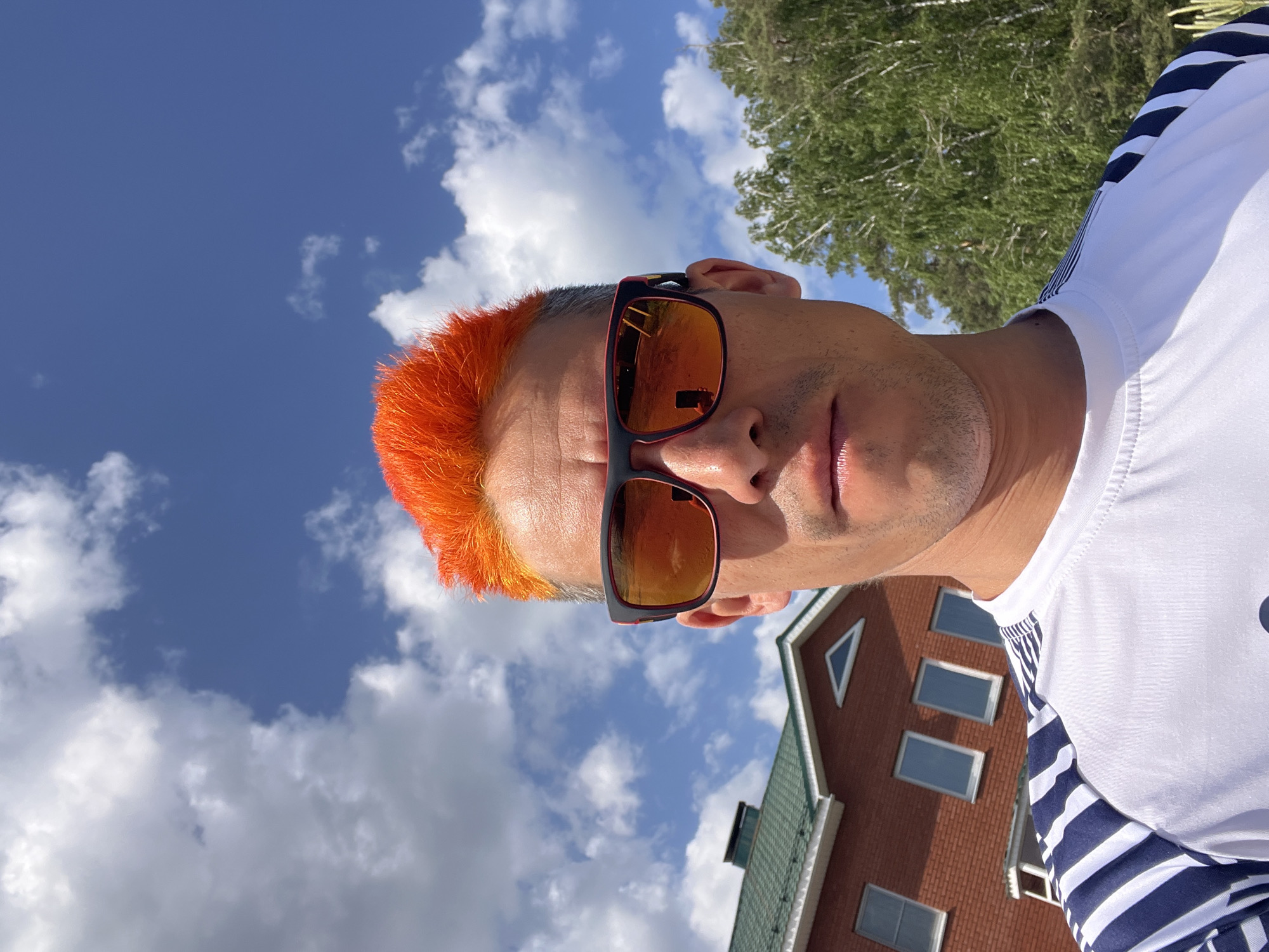 A selfie shows a man with bright orange hair and sunglasses against a backdrop of a partly cloudy sky, a tree, and part of a brick building. The man is wearing a white shirt with blue stripes on the sleeves. The building appears to have a green roof and several windows. The exact location in the city of Chaikovsky, Russia, is not discernable from the image.