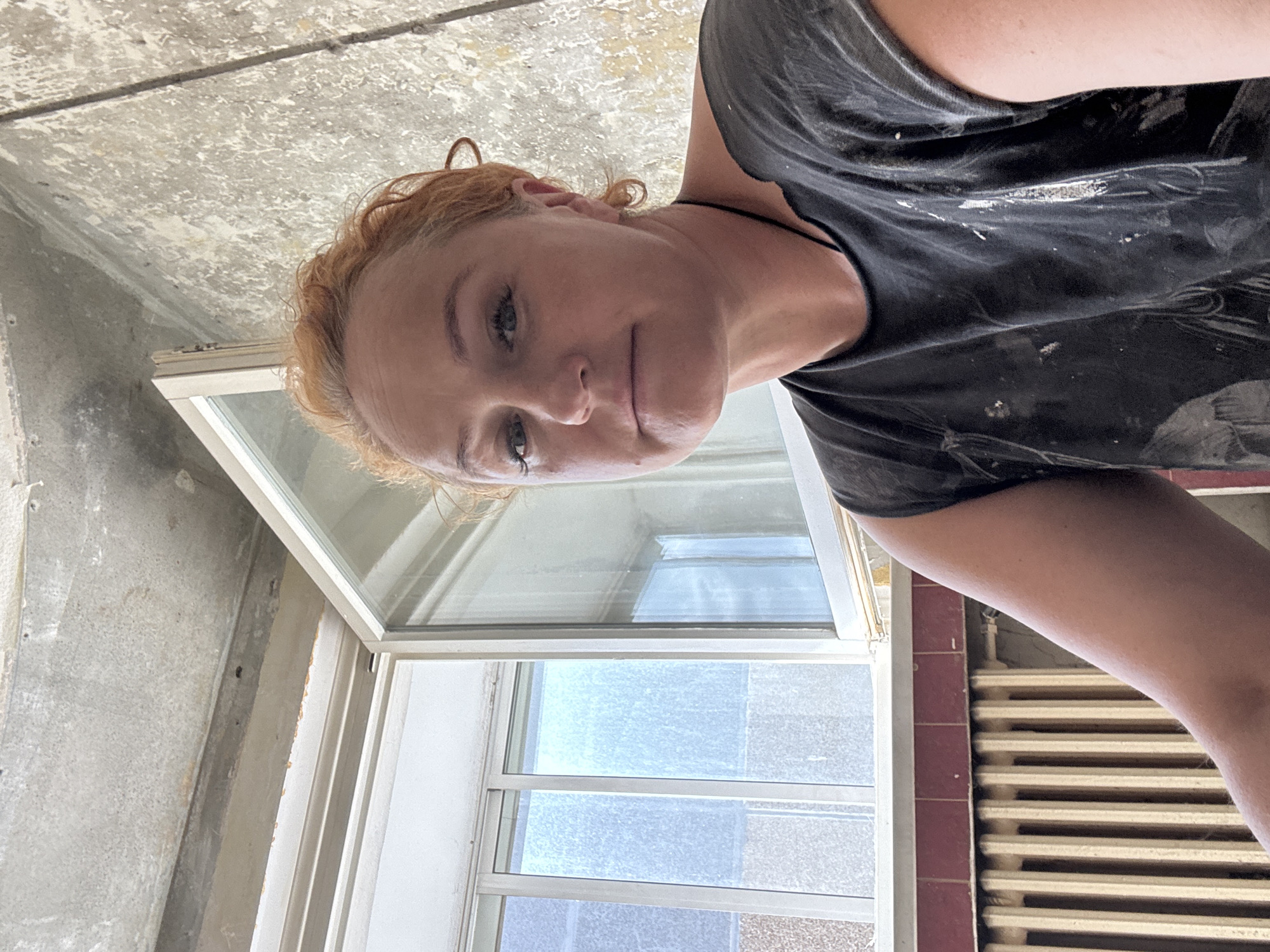 A woman with auburn hair is pictured indoors, in what appears to be a room undergoing renovation or construction. She is wearing a dark, possibly paint-splattered t-shirt. Behind her, a window is partially open, revealing a view of a building with multiple windows. A radiator is visible below the window. The ceiling appears to be unfinished concrete. The scene is likely in the city of Chaikovsky, Russia, given the context.