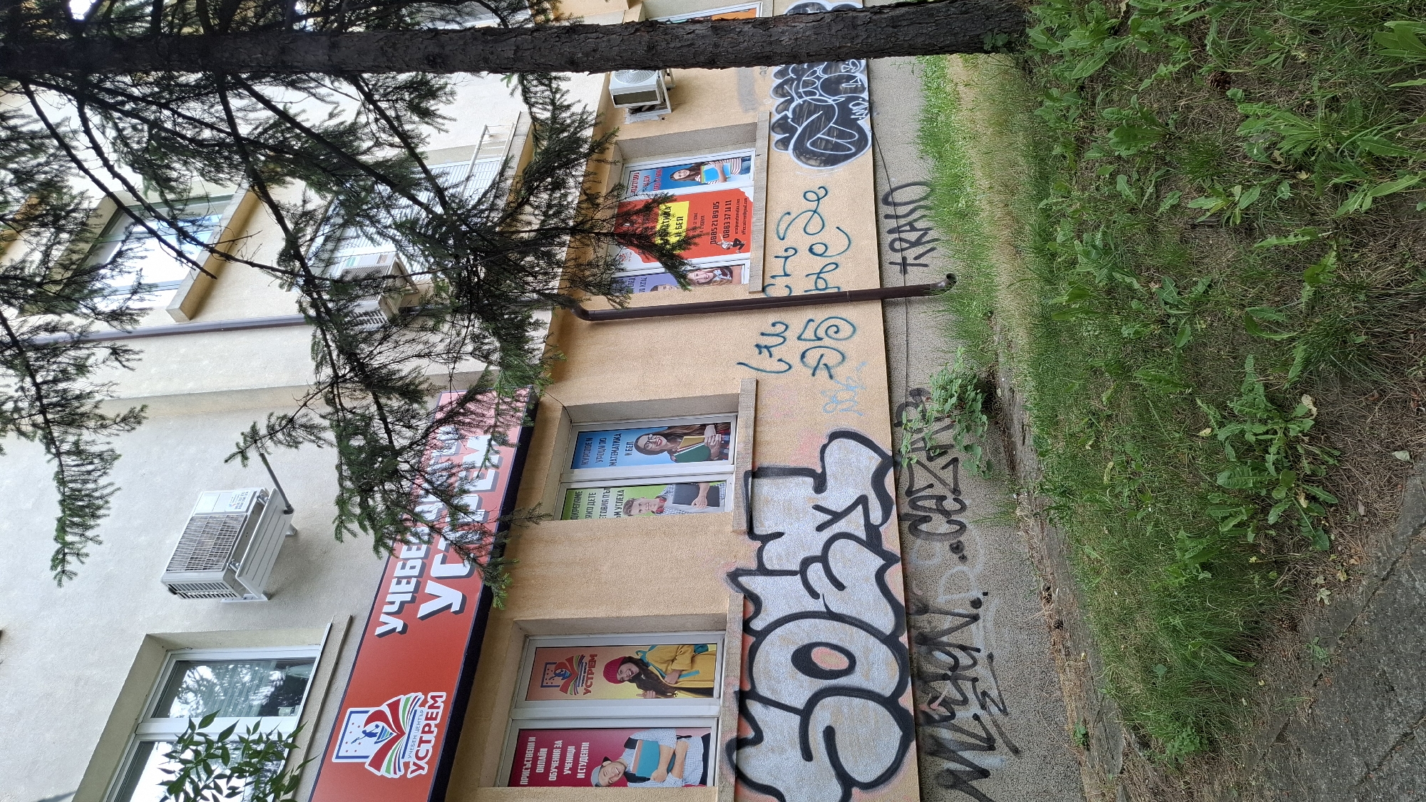 This media file shows a building exterior in what appears to be an urban setting, likely Sofia, Bulgaria. The building has multiple windows, some of which display advertisements for an educational center called "УСТРЕМ" (Ustream). These advertisements feature images of young people, presumably students, and text promoting various learning programs, including lessons in mathematics and Bulgarian language. One prominent advertisement on a red banner also displays the center's name.
The building's facade is a light tan color, and there is a drainpipe visible. An air conditioning unit is mounted on the wall above one of the windows. The scene is partially obscured by the branches and trunk of a large tree in the foreground, suggesting the building is situated along a street or sidewalk bordered by greenery.
Graffiti is visible on the building's wall and on the adjacent concrete pavement, adding a layer of urban character to the scene. The graffiti includes both stylized lettering and more abstract designs. Adjacent to the building and sidewalk, there is an area of overgrown grass and weeds. The lighting suggests it is daytime, but the exact time of day and weather conditions are not explicitly determinable from the image. The overall impression is one of a somewhat weathered, functional building in an everyday urban environment.