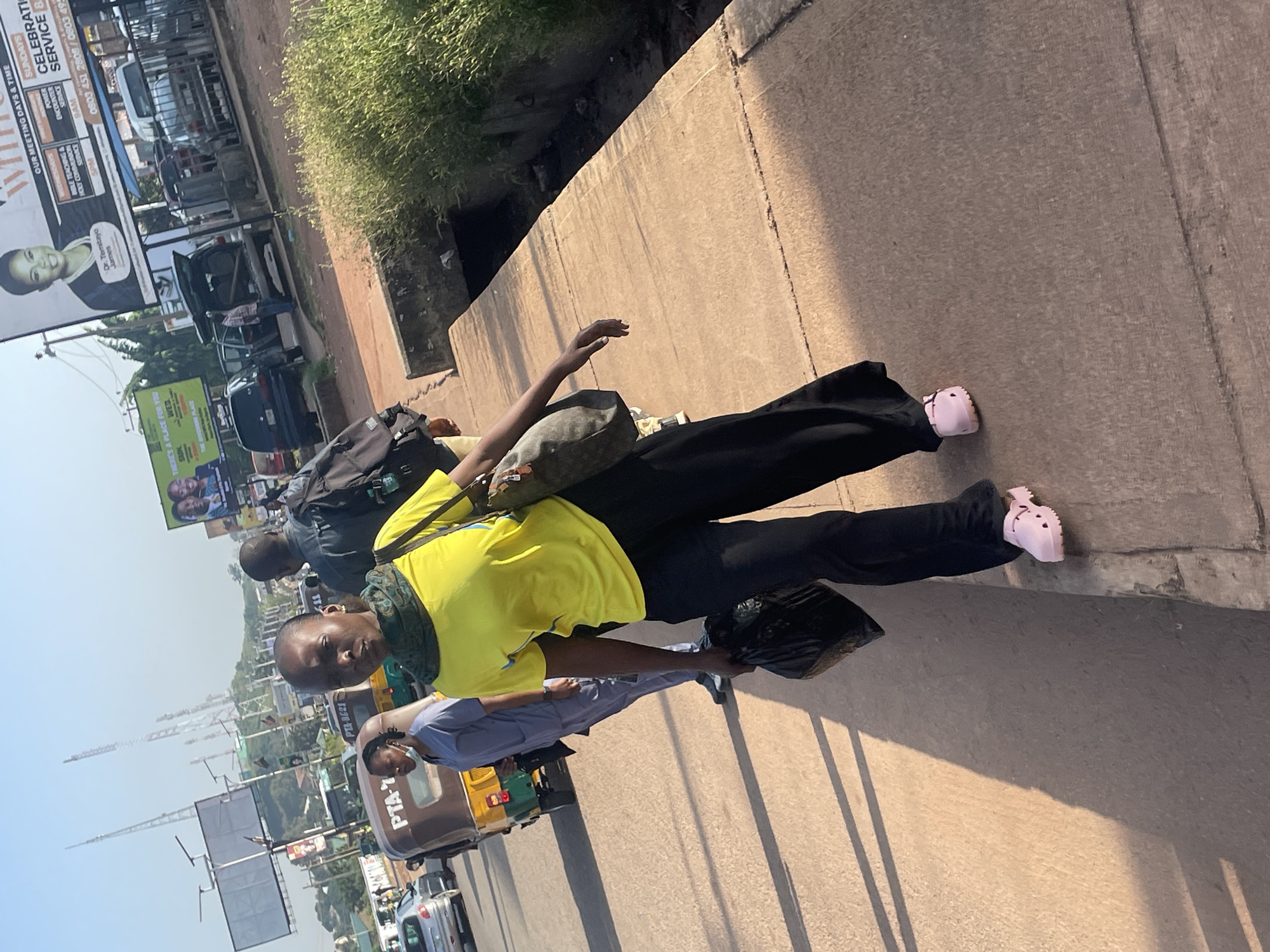 A young Black woman, wearing a bright yellow t-shirt, black pants, and pink Croc-style shoes, walks along a paved walkway in Jos, Nigeria. She carries a black plastic bag in her left hand and a brown handbag on her right shoulder. A green and black scarf is wrapped around her neck. Behind her, another Black person, wearing a blue medical scrub suit, walks in the same direction. Further behind them, a man in a dark shirt and backpack is visible. In the background, several vehicles, including a yellow and brown tuk-tuk, cars, and trucks, are on a busy street. Billboards are also visible in the distance, one featuring portraits of two men and text about meeting days and times, suggesting a religious or public event. The environment appears to be an urban street scene with overhead power lines and communications towers in the background. The ground is a mix of concrete pavement and a drainage ditch filled with green vegetation. The lighting suggests it is daytime with a low sun angle, casting long shadows.