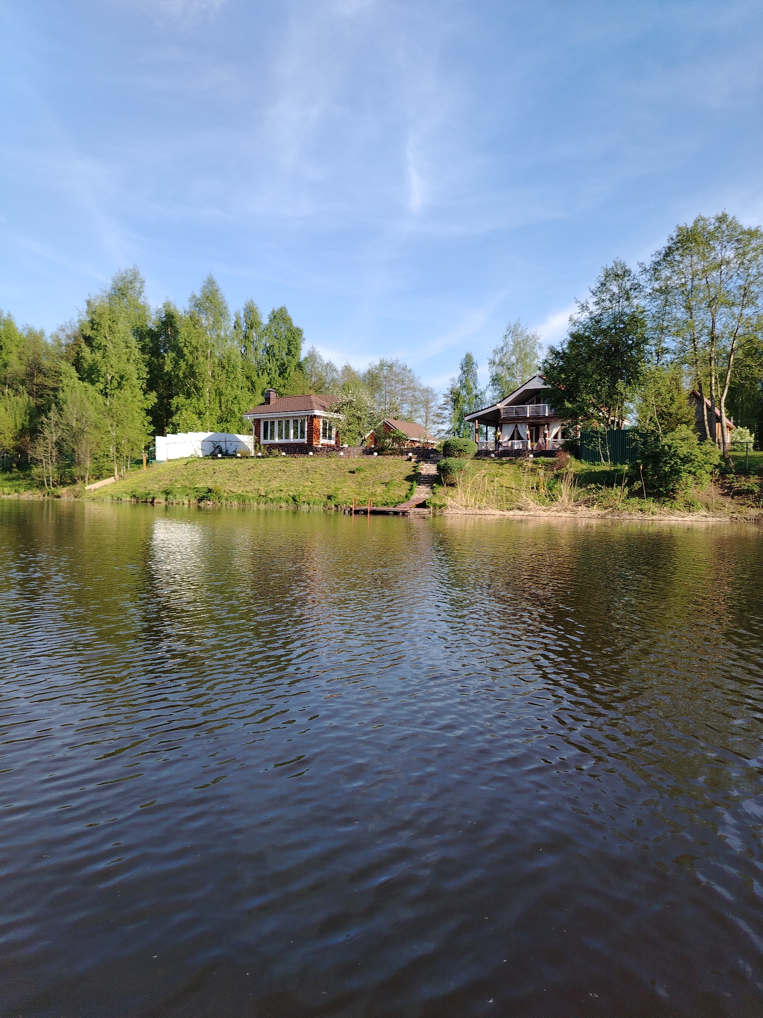 The image depicts a calm body of water, possibly a lake or wide river, with wooded banks and two buildings visible along the shoreline. The water is dark with visible ripples and reflections of the sky and shoreline.
The left bank features a small building with a red brick base and a brown roof, surrounded by trees. Further back, a white fence is visible.
On the right bank is a larger, two-story house with a brown roof and balcony. Trees and shrubs surround the house. A small wooden dock extends into the water near the house. The sky is blue with some scattered clouds.
The location is likely Stroitel', Russia, based on the image's context. No people are visible in the frame.