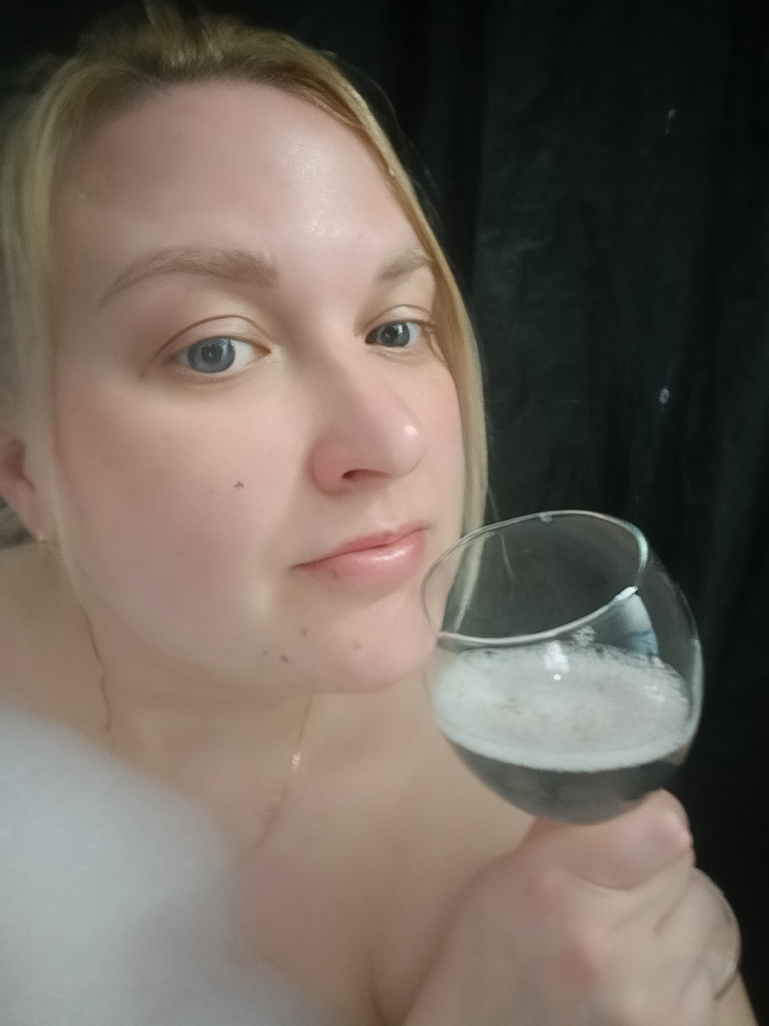 A blonde woman is holding a wine glass filled with bubbly liquid. The background is dark. The location and city cannot be determined.