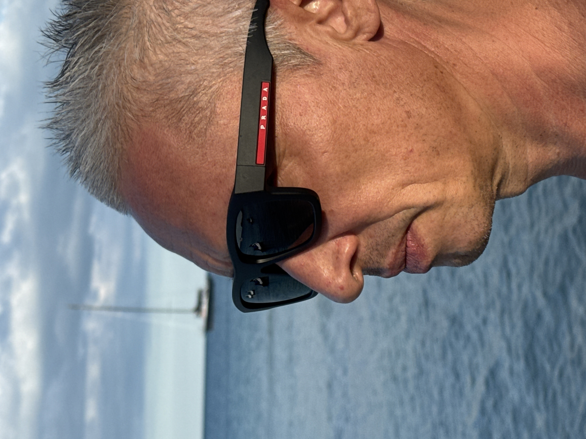 The image depicts a close-up of a person's head and upper shoulder, angled slightly downwards. The individual has short, grey hair and is wearing black Prada sunglasses. The background shows a body of water, likely the sea, with what appears to be a sailboat in the distance. Based on the location information provided, it is likely this photo was taken in or near Baan Chaweng, Thailand, but there is no evidence in the image to confirm this.