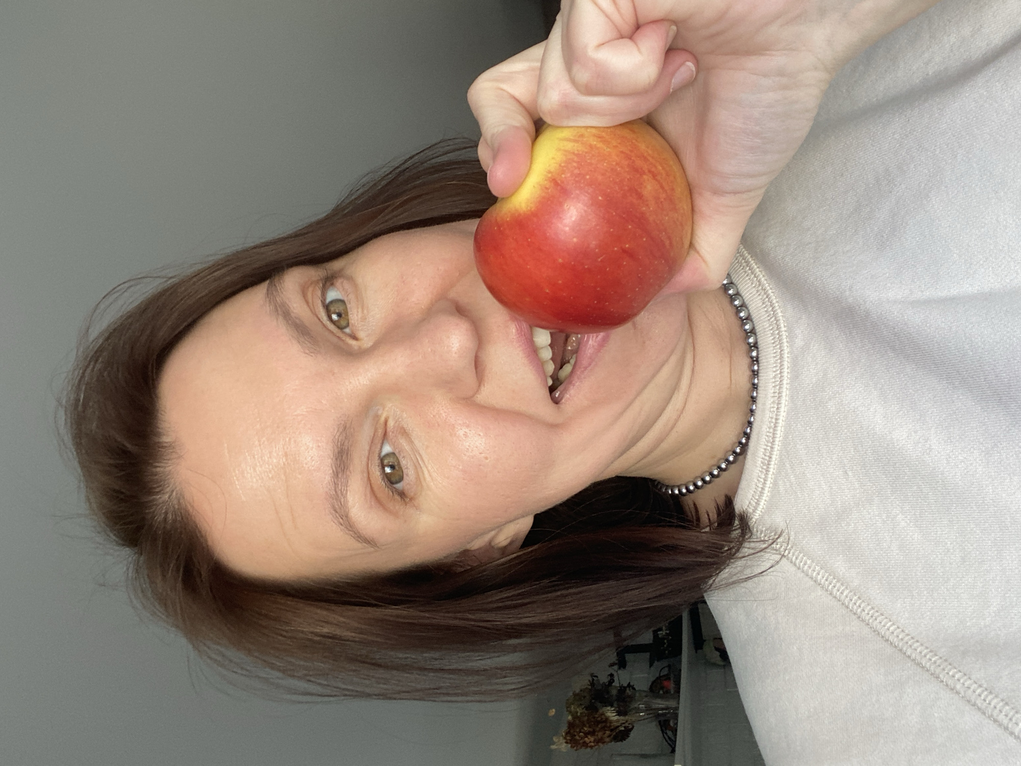 A woman is holding an apple to her mouth, appearing as if she is about to take a bite. The apple is red with some yellow hues. The woman has fair skin, brown hair, and green eyes. She is wearing a light-colored sweatshirt and a beaded necklace. The background is a plain grey wall. The location is unknown, but the query included Chaikovsky, Russia.
