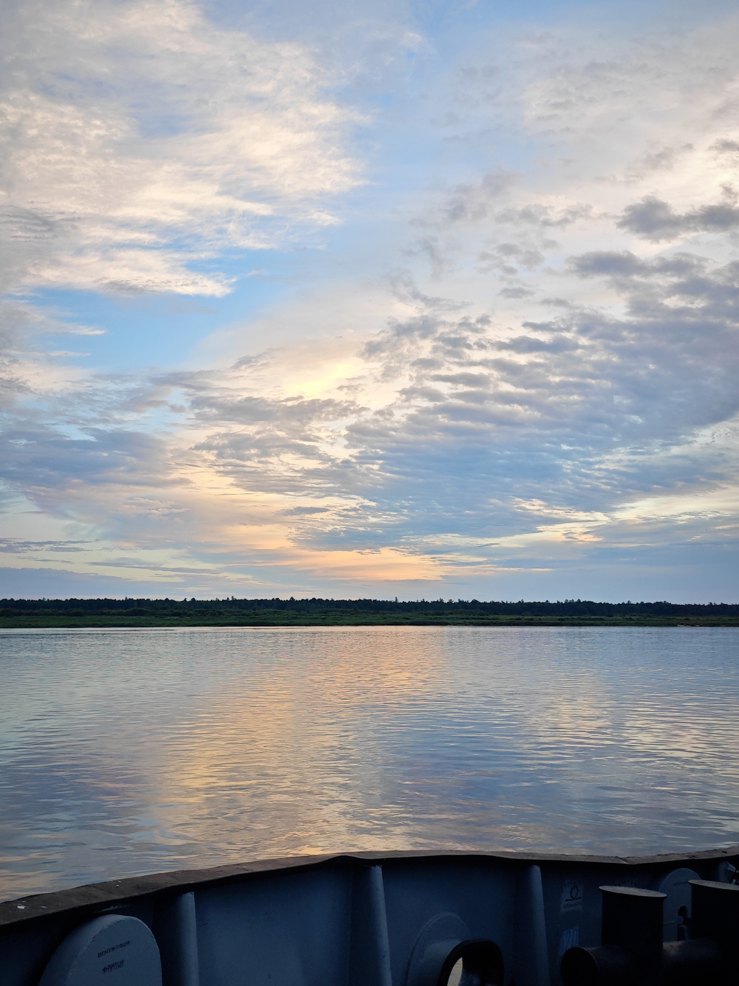 The image depicts a serene river scene at dusk, likely captured from the deck of a boat. The sky is a canvas of soft pastel colors, with streaks of orange, yellow, and pale pink mingling with shades of blue and grey. Wispy clouds are scattered across the sky, reflecting the fading sunlight on the calm water.
The river occupies the majority of the frame, its surface a mirror to the sky, creating a beautiful interplay of light and color. In the distance, a thin strip of dark green vegetation, possibly a treeline, marks the horizon. The foreground shows the edge of a boat, with visible metal structures and a life preserver, indicating the vantage point.
The overall mood is peaceful and tranquil, evoking a sense of calm as day transitions into night. The time of day is clearly evening, given the soft, diffused light and the colors in the sky. The weather appears clear and mild.
There is some visible text on the white circular object in the foreground, possibly a label for the life preserver. The text is in Russian and appears to include Cyrillic characters, but it is not clear enough to be fully deciphered. The location context provided is Nikulino, Russia.