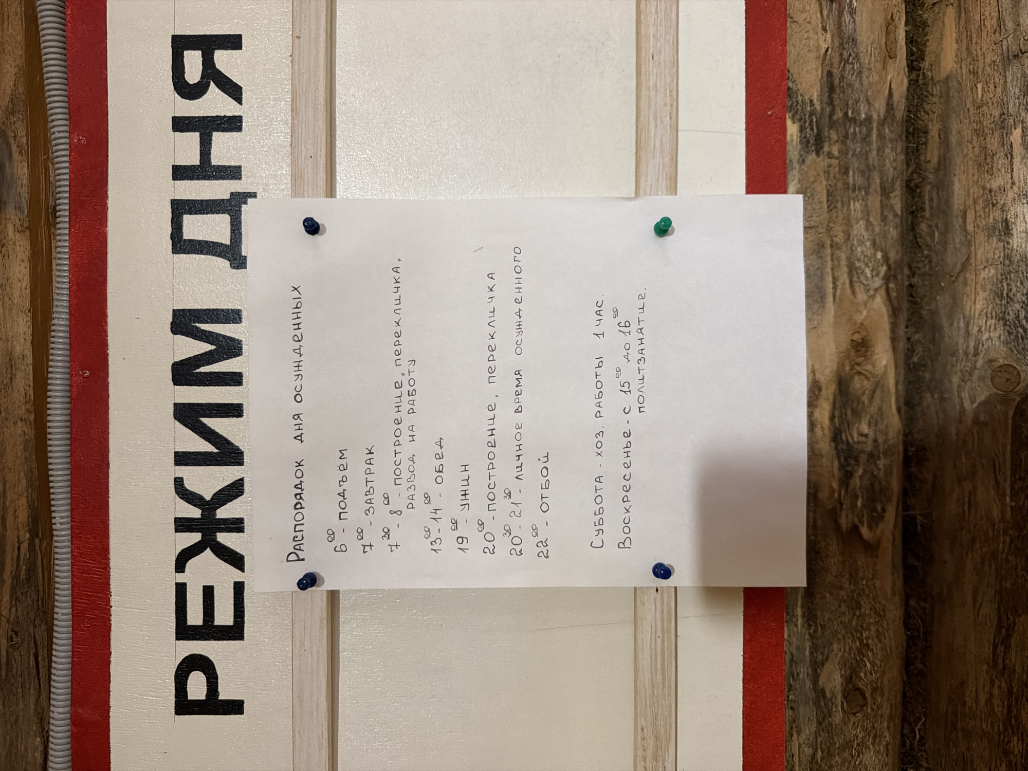 The image shows a handwritten schedule pinned to a white board with a red stripe at the top. The schedule is written in Russian. Large black letters at the top of the board spell out "РЕЖИМ ДНЯ," which translates to "Daily Schedule." Below this, a smaller heading reads "Распорядок дня осужденных," meaning "Schedule for the condemned."
The schedule details the daily routine:
* **6:00** - подъём (wake-up)
* **7:00** - ЗАВТРАК (breakfast)
* **7:30 - 8:00** - ПОСТРОЕНИЕ, ПЕРЕКЛИЧКА, РАЗВОД НА РАБОТУ (assembly, roll call, dispatch to work)
* **13:00 - 14:00** - ОБЕД (lunch)
* **19:00** - УЖИН (dinner)
* **20:00** - ПОСТРОЕНИЕ, ПЕРЕКЛИЧКА (assembly, roll call)
* **20:30 - 21:00** - ЛИЧНОЕ ВРЕМЯ ОСУЖДЕННОГО (personal time for the condemned)
* **22:00** - ОТБОЙ (lights out)
The schedule also includes weekend hours:
* **Суббота** - хоз. работы 1 час. (Saturday - 1 hour of housekeeping duties.)
* **Воскресенье** - с 15:00 до 16:00 - ползанятие. (Sunday - from 15:00 to 16:00 - half-session, likely referring to a period of activity or instruction.)
The overall environment appears to be utilitarian, with the wooden background suggesting an interior space that might be a facility or institution. The presence of pushpins indicates the schedule is intentionally displayed for viewing. The context implies this is a schedule within a correctional facility or a similar institution for individuals who are "condemned" or "convicted."
There are no people visible in the image. The scene is static. The location is specified as Ust'-Pyzas, Russia, which is a known location for correctional facilities.
