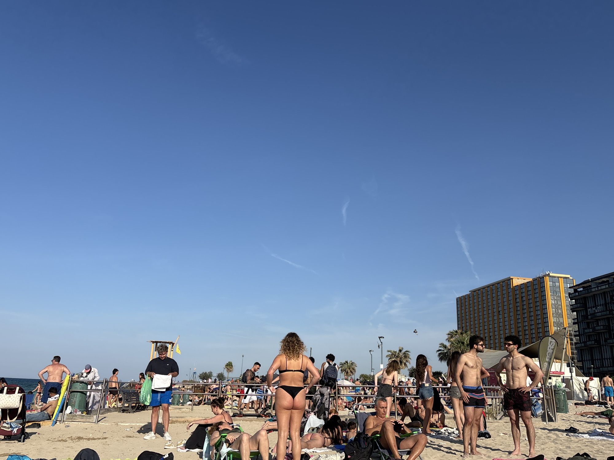 The image depicts a bustling urban beach scene under a clear, bright blue sky. Numerous people, primarily adults, are visible across the sandy expanse, enjoying a sunny day. Many are dressed in swimwear, such as bikinis and swim shorts, indicative of warm weather.
In the foreground and mid-ground, individuals are seen engaging in various leisure activities: sunbathing on towels, relaxing in beach chairs, standing and conversing, or walking around. On the left, the sea is visible in the distance, and a yellow wooden lifeguard stand can be seen further back. In the mid-left, a man in a black t-shirt and blue shorts stands holding papers and a green bag near a public green trash bin. Centrally, a woman with curly hair is viewed from behind, wearing a black bikini. Towards the right, two shirtless men in swim shorts stand facing each other, engaged in conversation.
The background features several tall buildings, most notably a prominent yellow-orange high-rise with many windows, suggesting a hotel or apartment complex. Other darker, modern buildings are also visible, along with several palm trees scattered throughout the scene. The weather is sunny with a clear blue sky, showing only faint contrails, and the strong sunlight indicates it is daytime, likely mid-day or afternoon. The overall atmosphere is lively and recreational. No discernible text is visible on any objects or signs.