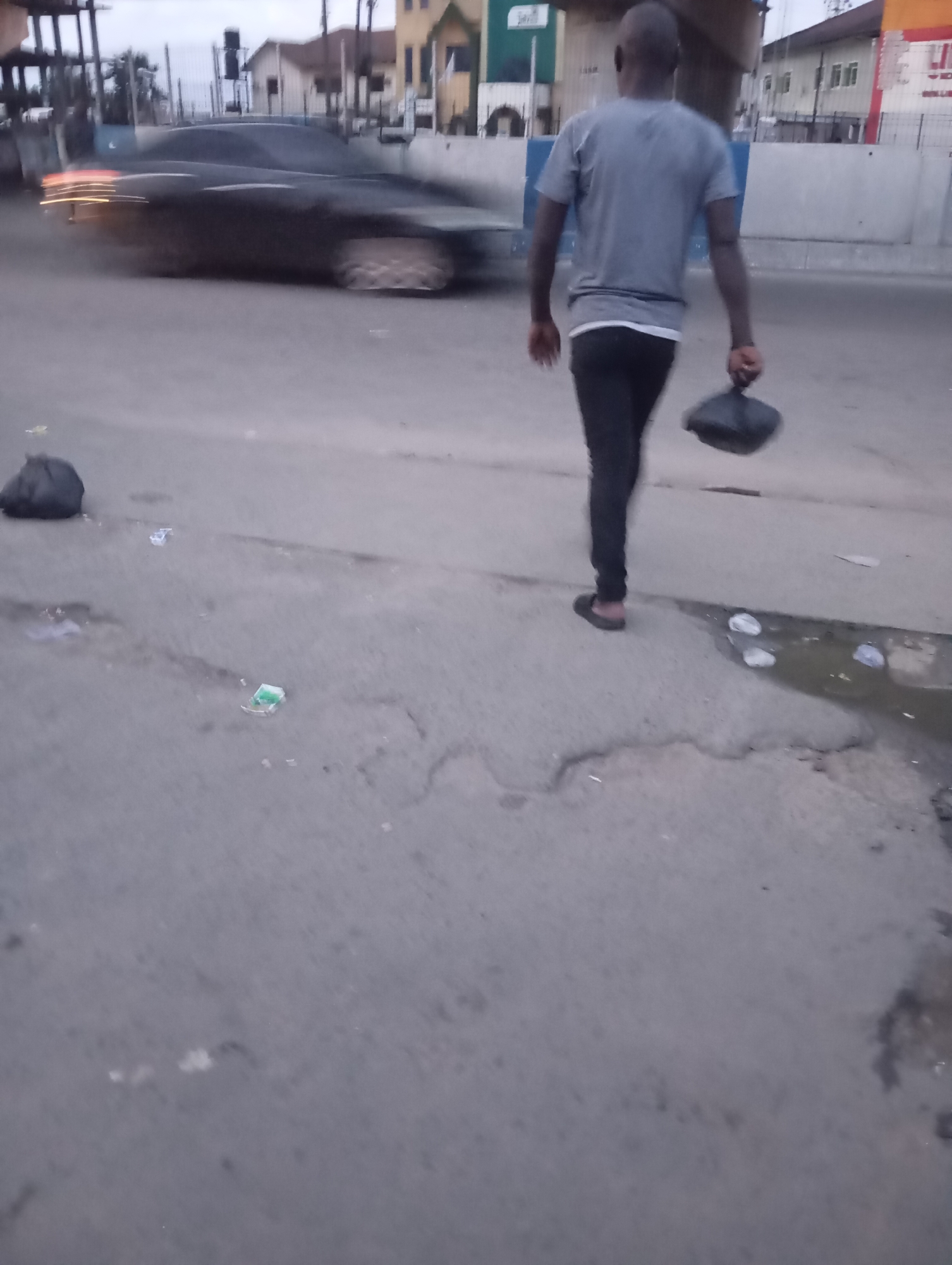 In Port Harcourt, Nigeria, an out-of-focus, medium shot shows a person walking away from the camera on a paved surface. The individual, a bald male wearing a grey t-shirt and black pants, is carrying a black plastic bag in his right hand. To the left of the frame, a black garbage bag is visible on the ground. In the background, a dark-colored car with blurred red taillights is in motion, suggesting traffic. Buildings and a water tower are also discernible in the distance, indicating an urban environment. The ground is littered with small pieces of debris.