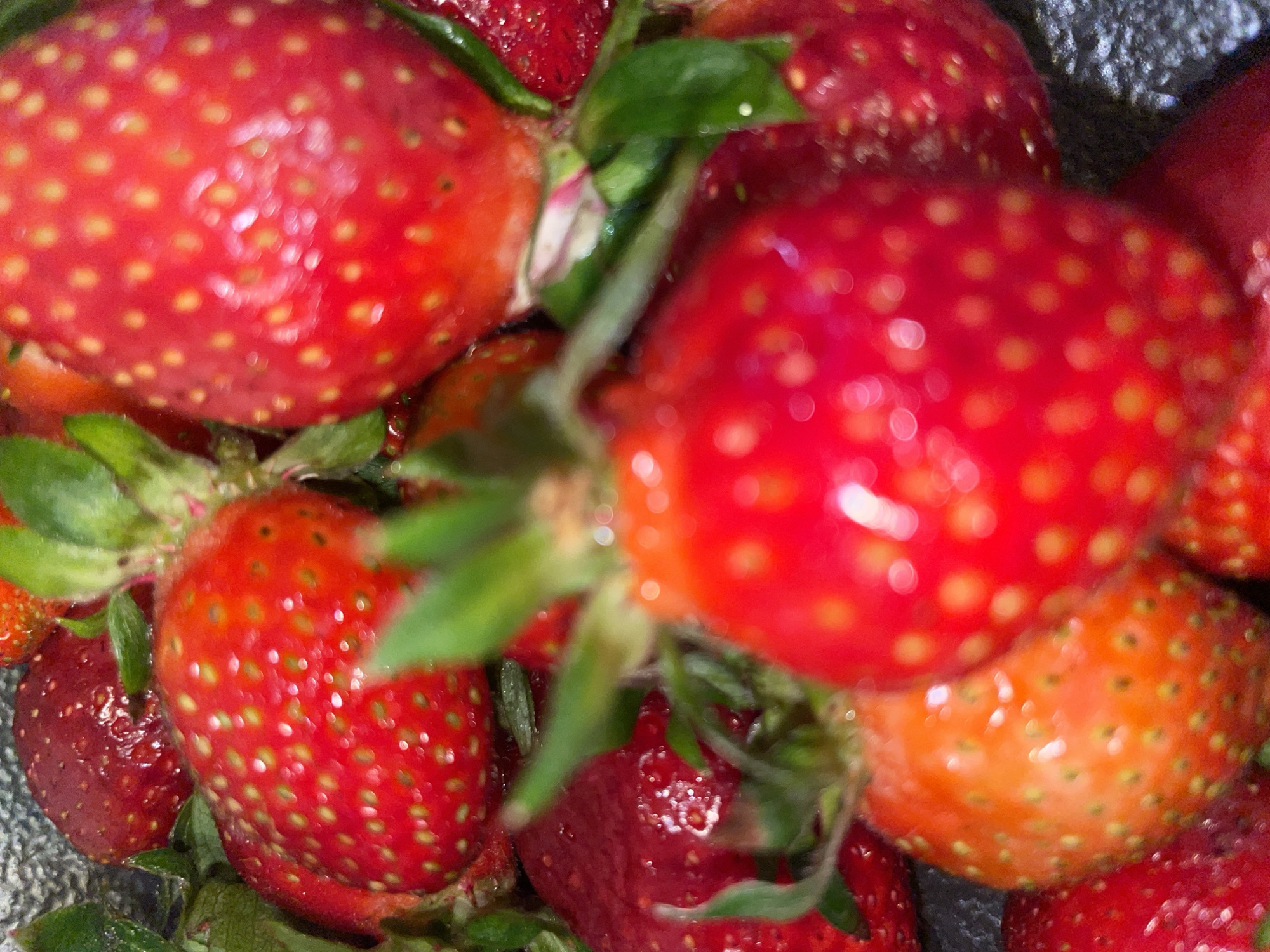 A close-up, slightly out-of-focus photograph shows a cluster of fresh strawberries. The strawberries vary in ripeness, with some appearing bright red and others exhibiting orange or darker red hues. Small green leaves are visible amidst the strawberries. The strawberries are covered in small, yellow seeds. The background is a gray, textured surface. The location is not specified in the image.