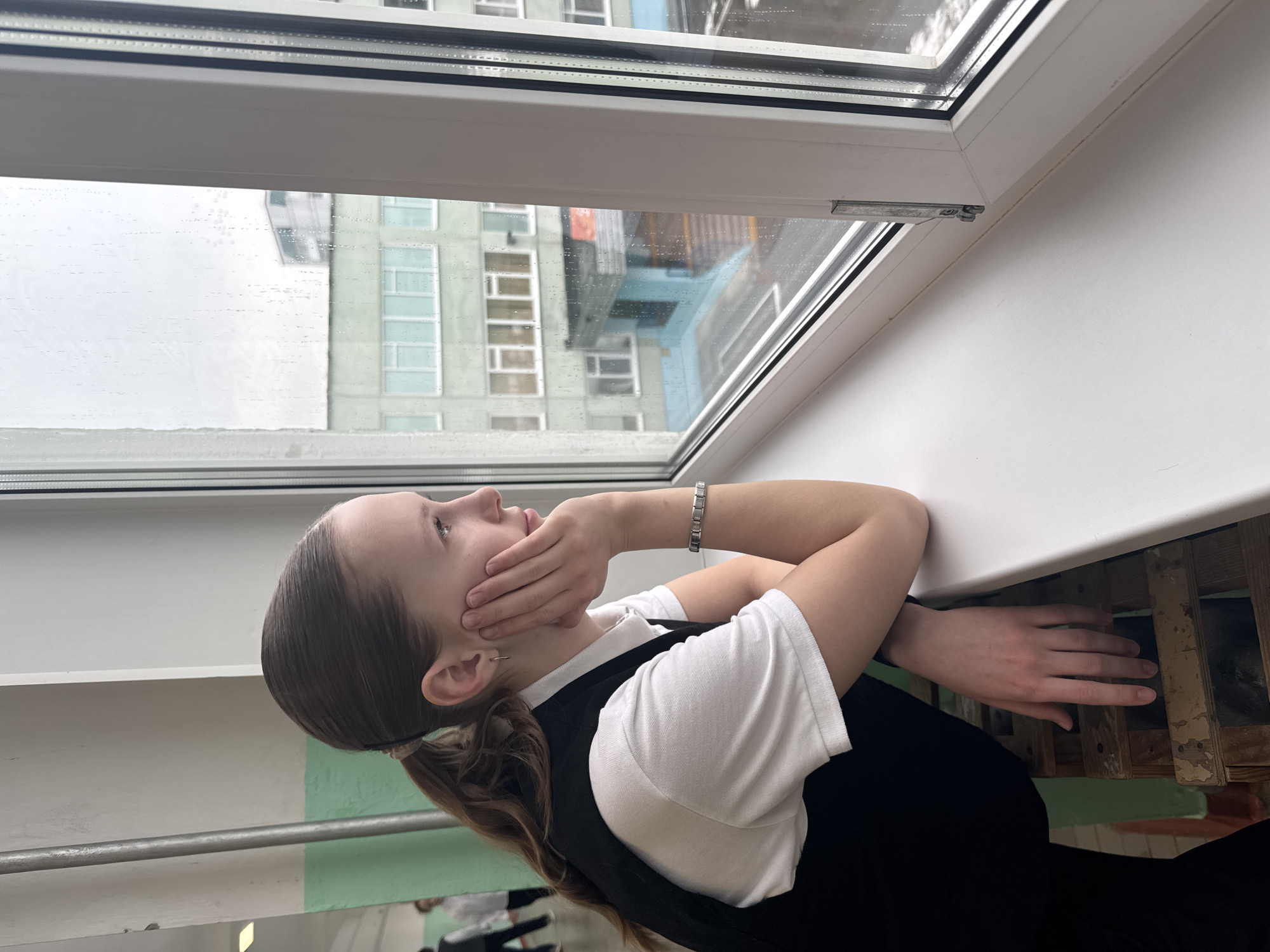 A young woman, possibly a school student, is resting her head on her hand, looking up and out of an open window. She is wearing a white shirt with a black vest or jumper, and a silver bracelet. Her brown hair is tied back in a ponytail. The window overlooks a cityscape, with buildings visible through the rain-streaked glass. The window frame is white, and the sill appears to be under renovation. The location is most likely Chaikovsky, Russia, based on the buildings that are seen. The surrounding environment appears to be inside a building, near a wall.