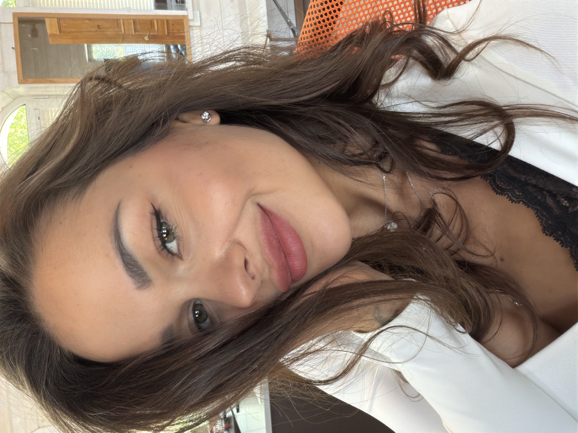 A selfie is taken of a woman from the chest up. She has long, brown hair that is styled loosely, and her eyes are green. She is wearing a white blazer over a black lace top. Her lips are painted with a pink lipstick, and she has a light dusting of blush on her cheeks. A delicate silver necklace with a small pendant rests on her collarbone. A small, sparkly earring is visible on her left ear. The background appears to be an indoor setting, possibly an office or living space. There is a window with natural light coming through, and a reflection in what looks like a mirror shows a doorway and part of a room. To the right, an orange chair with a mesh-like texture is partially visible. The image is well-lit, focusing on the woman's face and upper body.