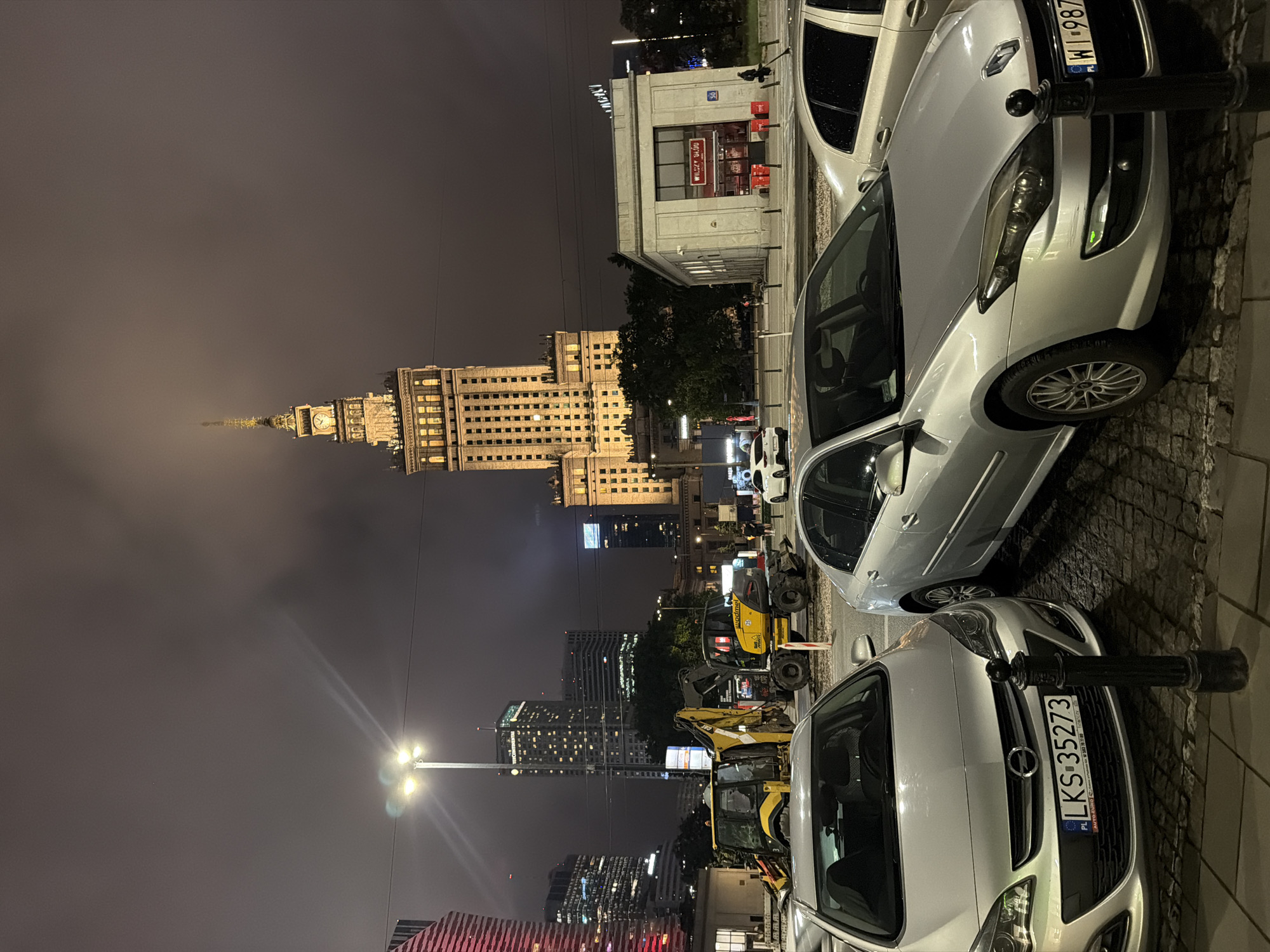 In an urban nighttime scene, a large, illuminated building, resembling the Palace of Culture and Science in Warsaw, dominates the background. The sky is heavily overcast, obscuring the upper portions of the building and creating a muted, hazy atmosphere. In the foreground, several silver cars are parked on a cobblestone street. A black bollard stands in front of the closest car, an Opel with a Polish license plate that reads "LKS 35273". To the right of the Opel, a silver Renault Laguna is parked, its license plate obscured by a black bollard. In the middle ground, construction equipment, including a yellow excavator and a backhoe loader, is present on the street, indicating ongoing road work. Adjacent to the construction area, a small, light-colored building with red signage is visible. Other modern, multi-story buildings with illuminated windows are scattered throughout the cityscape. A streetlamp with three lights is positioned in the middle left of the frame. A few pedestrians are visible in the distance, walking near the illuminated buildings and construction site. The overall impression is of a busy city at night, with a mix of architectural styles and ongoing urban development.