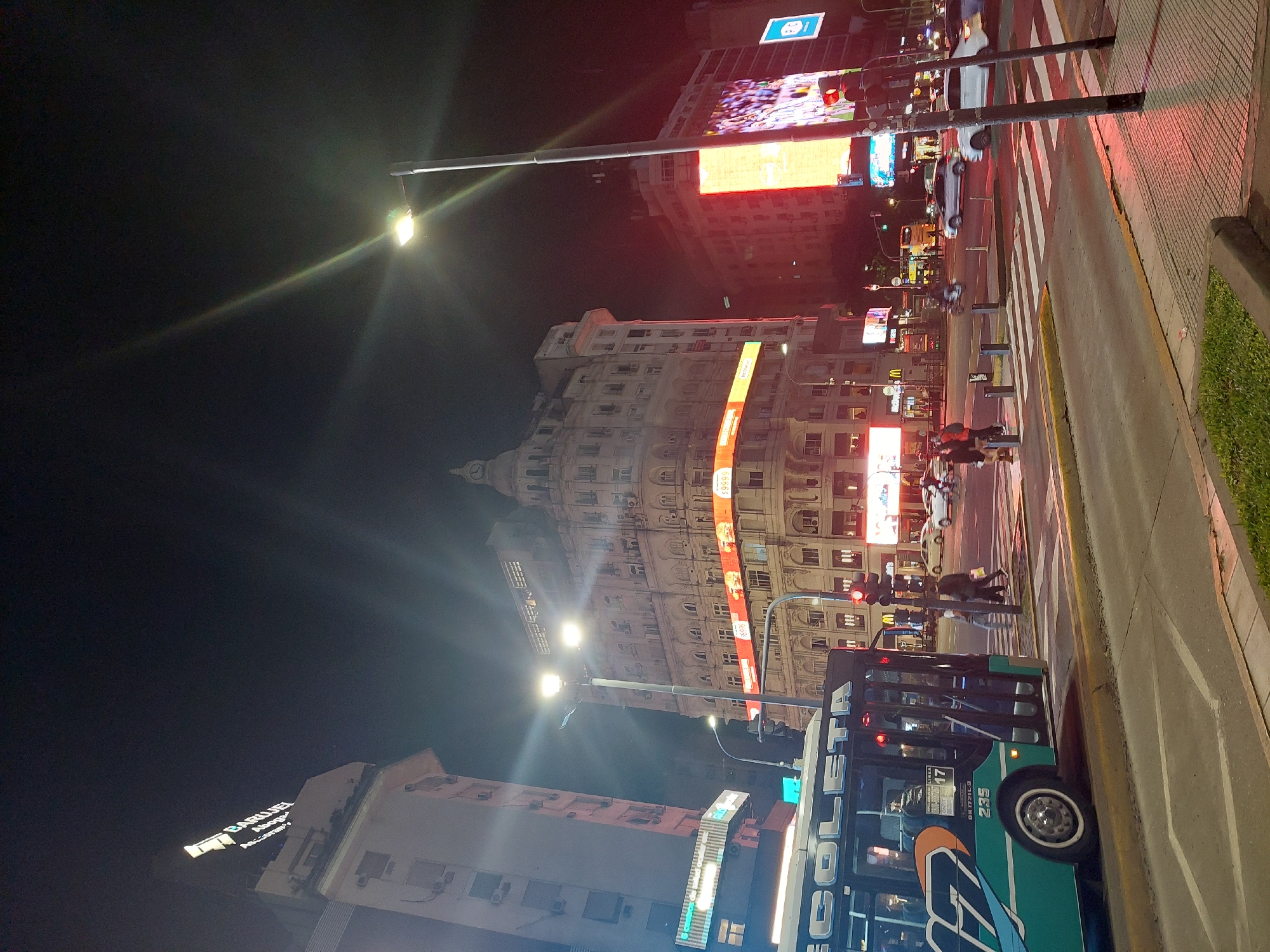 The image captures a lively urban street scene at night in Buenos Aires, Argentina. The perspective is from the sidewalk, looking down a bustling avenue lined with multi-story buildings. The sky is dark, indicating nighttime, and the area is brightly illuminated by numerous artificial light sources.
In the foreground, a sidewalk with a narrow strip of green grass borders a multi-lane street. A prominent pedestrian crosswalk with thick white stripes is visible across the road. A green and blue city bus, marked "COLETA" and "LINEA 17", with the number "235" on its front, is partially visible on the right side of the road, appearing to be stopped or moving slowly.
Along the street, several cars are present, some moving, others potentially stopped at a traffic light, which appears red in the distance. Pedestrians can be seen walking or waiting on the sidewalk and near the crosswalk.
Tall buildings with classical architectural details dominate the background and midground, their facades lit up by streetlights and large digital billboards. One particularly ornate building features a dome-like structure at its top. Bright light rays emanate from streetlights positioned along the avenue, creating starburst effects in the night sky.
Multiple large digital screens display colorful advertisements and possibly news or sports content, adding to the dynamic ambiance. One partially visible sign on a building to the far left reads "BARQUEL Hogar y Accesorios". Other numbers like "10969" are visible on a red banner on a building. The overall impression is one of a vibrant city environment after dark, bustling with traffic and activity.