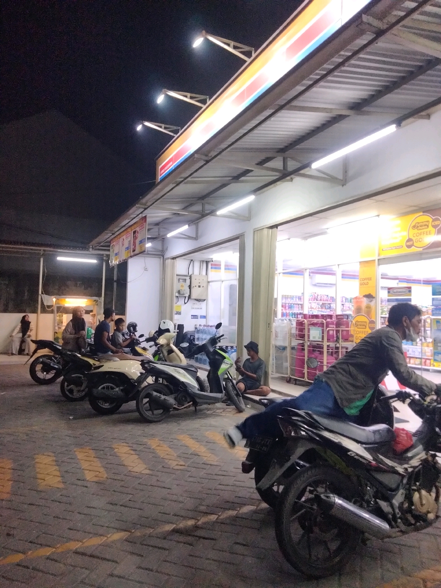 A group of people are gathered outside a convenience store at night. Several motorcycles are parked in front of the store. In the foreground, a man is sitting on a motorcycle, leaning back. He is wearing a dark jacket and blue jeans. Behind him, other people are seen standing and sitting near parked motorcycles. The convenience store has bright interior lighting and signage. The ground is paved with interlocking bricks and marked with yellow lines. The overall scene appears to be a casual gathering at a local store in Pisangan Jaya, Indonesia.