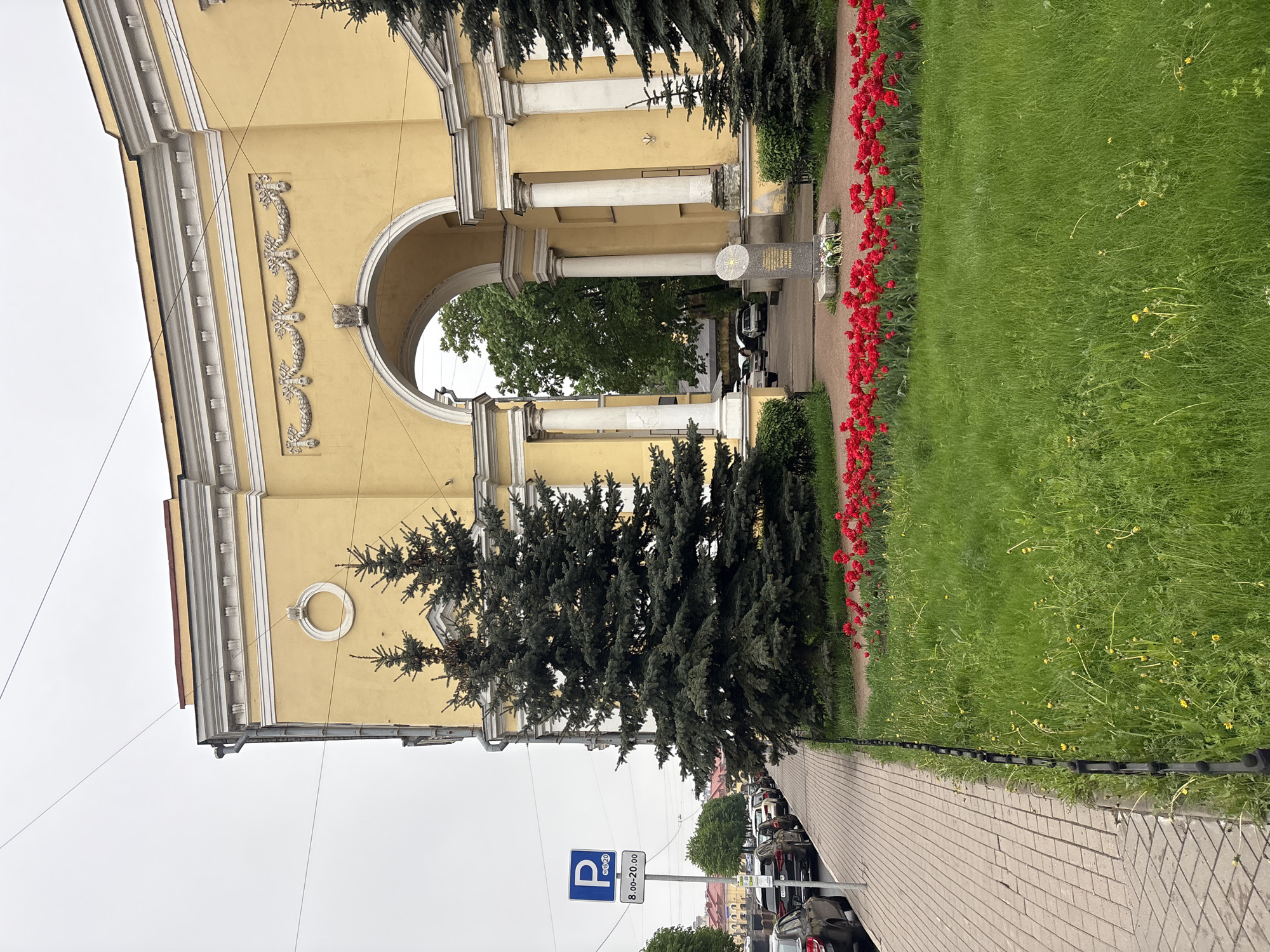 The image depicts the Pskov Gates in Saint Petersburg, Russia. The gates are constructed of yellow stucco and feature a large archway supported by white columns. There are decorative elements on the facade. A dark green spruce tree stands in front of the structure on the left. A pathway of light brown pavers runs along the right edge of the image. A border of red tulips lines the path, next to a grassy area. A parking sign is visible to the left of the spruce tree, indicating paid parking between 8:00-20:00. Several parked cars are along the street beyond the parking sign. The sky is overcast.