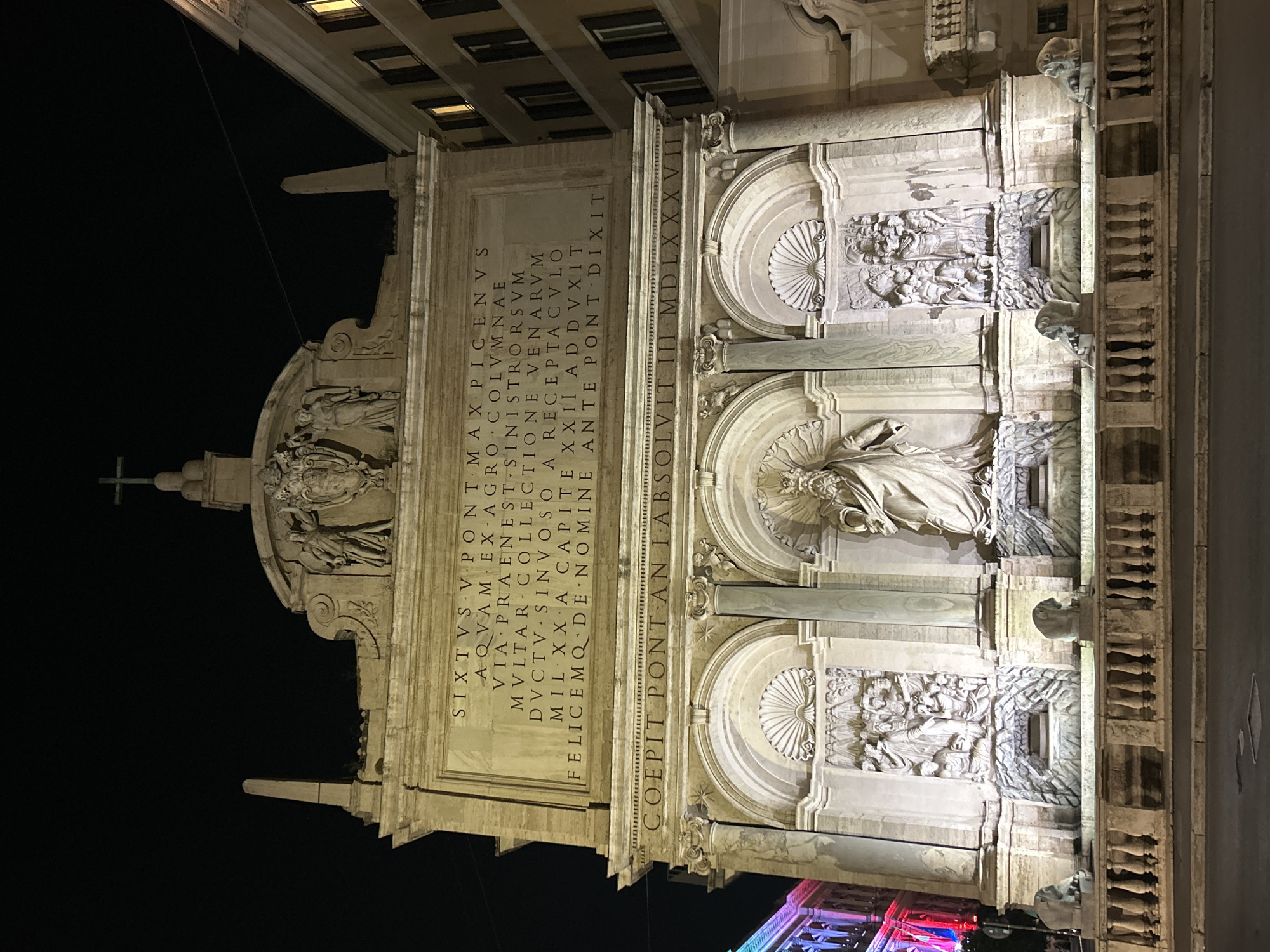 The image shows the Fontana dell'Acqua Felice in Rome, Italy, at night. The fountain is a large Baroque structure with multiple levels and sculpted figures. The main section of the fountain features a central niche with a statue of Moses, flanked by two smaller niches containing bas-reliefs. Above the niches, there is an inscription in Latin. The fountain is illuminated, casting shadows and highlighting the intricate details of the sculptures. The surrounding area appears to be a street with buildings on the right and a glimpse of a colorful building on the left.