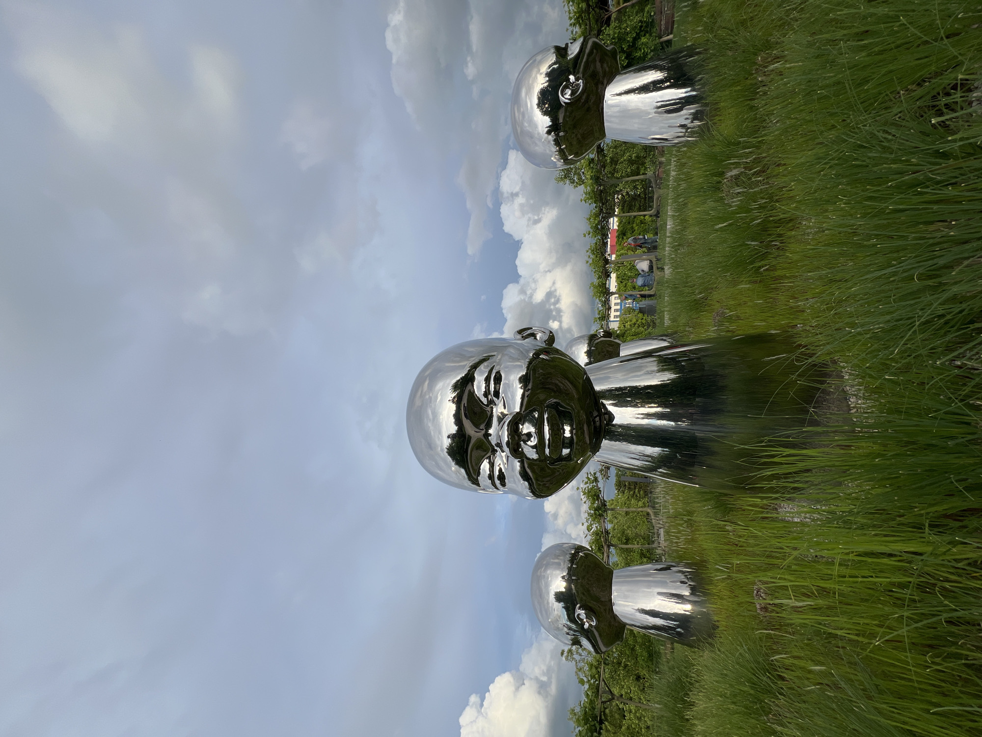 The image shows three large, chrome-plated sculptures of baby faces, set in a grassy area under a cloudy sky. The sculptures are arranged vertically in the image, with one large face in the middle and two smaller faces above and below it. The chrome surfaces of the sculptures reflect the sky and surrounding greenery. The scene appears to be a park or outdoor art installation. While I have difficulty determining the location, the sculptures are located in the city of Krasnodar, Russia. The background includes trees, and in the distance, there are hints of buildings and people. The overall impression is one of modern art in a natural setting.