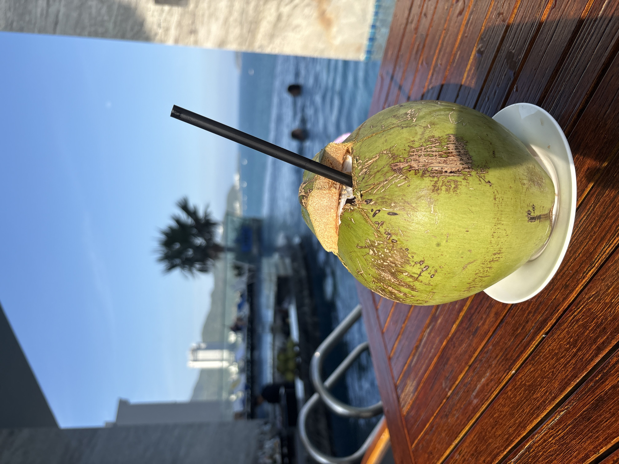 The image captures a coconut drink on a wooden table, likely near a swimming pool overlooking the ocean. The coconut, served on a white plate, has a black straw inserted. In the background, the ocean is visible along with some indistinct figures in the pool and on the shore. A palm tree and a building, potentially a hotel, are also present in the background. The location is presumed to be Nha Trang, Vietnam.