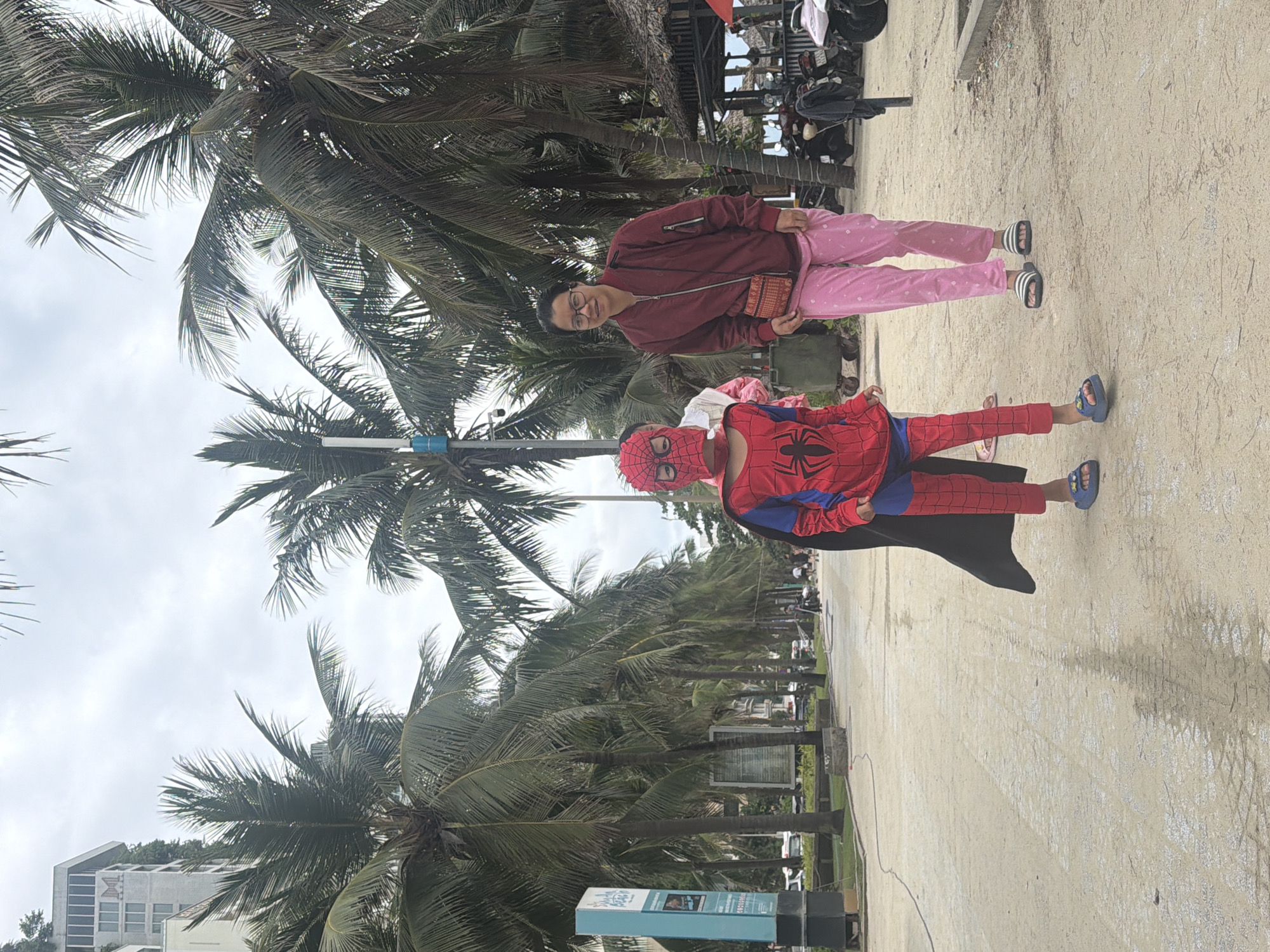 On a sunny day in Đà Nẵng, Vietnam, a woman wearing glasses, a maroon jacket, and pink pants stands next to a child dressed in a Spider-Man costume with a black cape. They are both standing on a sandy area next to a street lined with palm trees. In the background, there are buildings and vehicles.