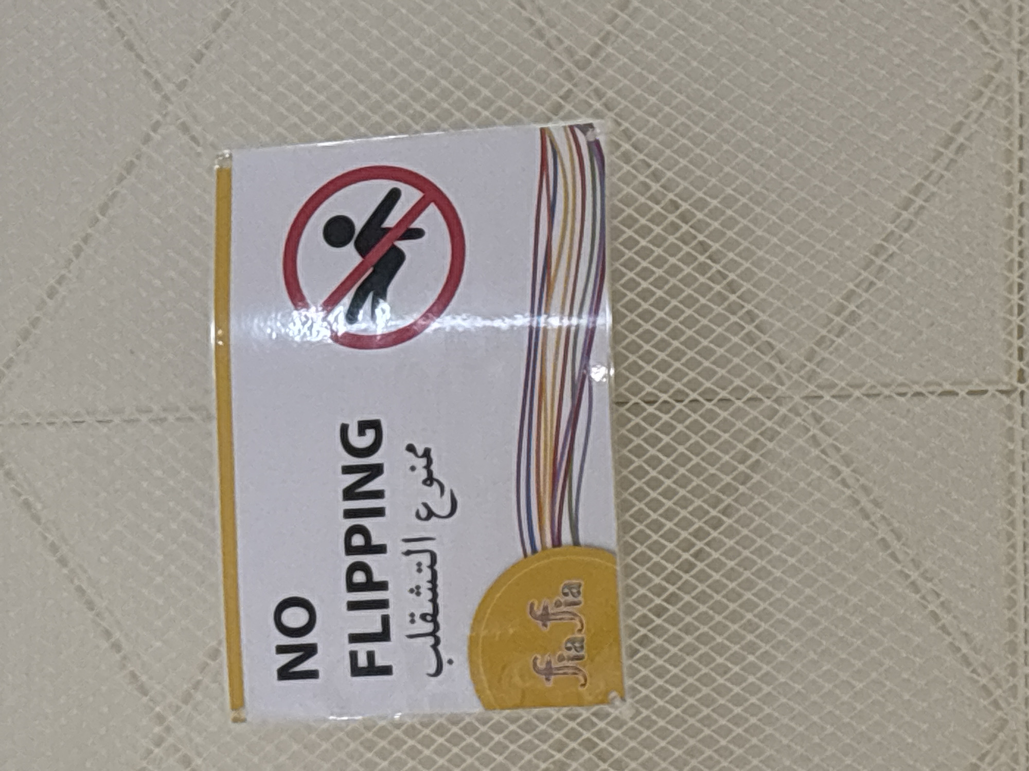 The image is a close-up, eye-level shot of a sign on a textured, light-colored surface, possibly a mattress or upholstered furniture. The sign has a yellow border at the top and appears to be made of laminate or plastic. It contains text in English and Arabic, along with a graphic symbol.
The English text reads "NO FLIPPING" in large, black, sans-serif font. Below this, in Arabic, it reads "ممنوع التشقلب" (mamnoua altashqlb), which also translates to "No Flipping". To the right of the text is a red circle with a diagonal line through it, indicating prohibition. Inside the circle is a black silhouette of a person in a dynamic pose, appearing to be falling or flipping.
In the bottom left corner of the sign, there is a yellow circular emblem with a stylized logo that appears to be "fia fia" written in cursive. Below this emblem, the sign features a band of colorful horizontal wavy lines in shades of red, orange, yellow, blue, and purple, suggesting a playful or decorative element.
The background surface has a diamond-patterned texture, common in upholstery or bedding. The lighting is bright and direct, creating some glare on the sign. The overall scene is clean and appears to be an indoor setting. There is no information within the image itself that indicates a specific location in Dubai, United Arab Emirates.