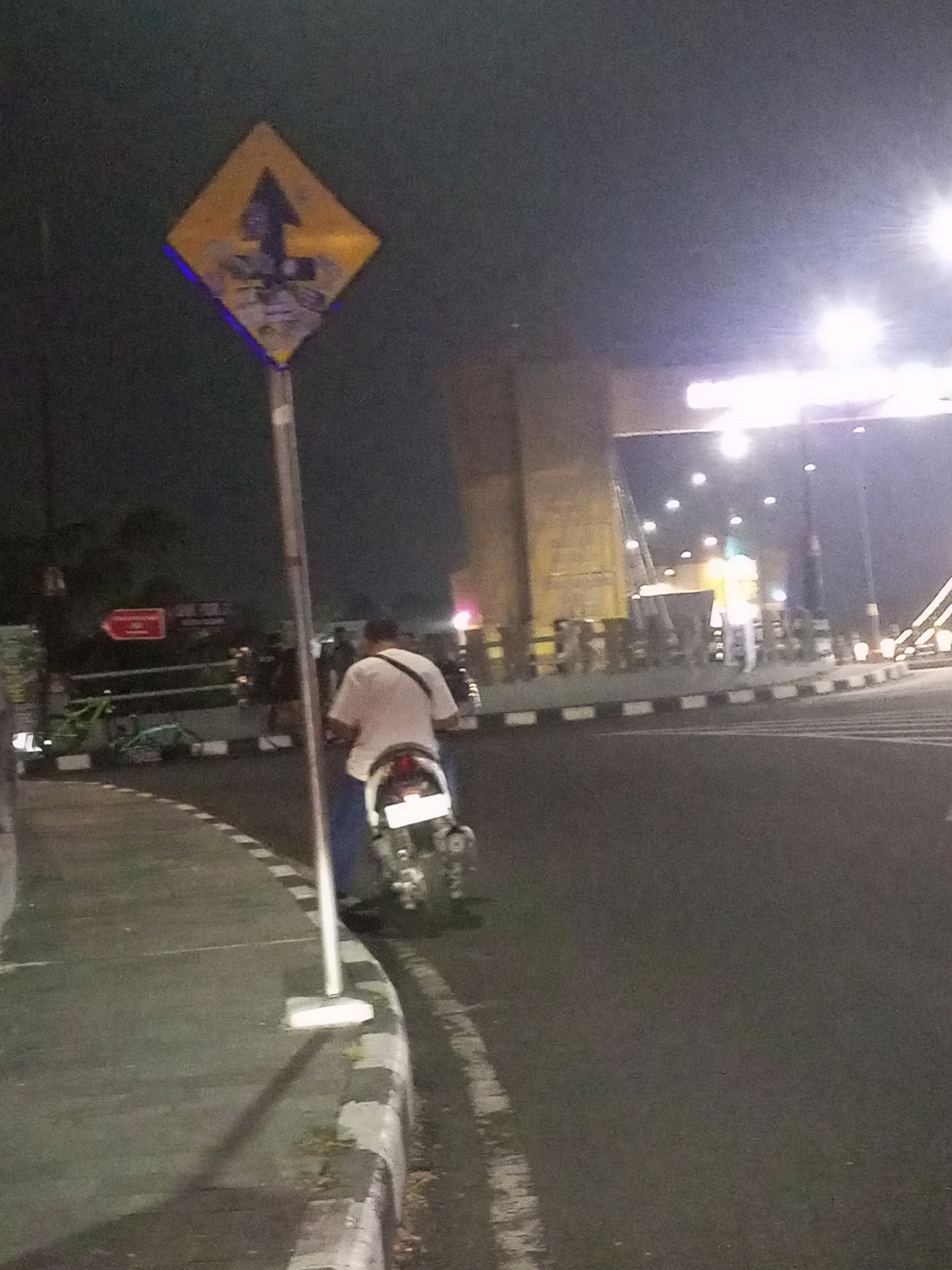 A person is riding a motorcycle at night on a street in Kediri City, Indonesia. The motorcycle has a white license plate. The rider is wearing a white t-shirt and blue pants and has a bag slung over their shoulder. A diamond-shaped yellow traffic sign with a blue border is visible on the left side of the road, indicating a turn or split in the road ahead. The background shows streetlights and a large building structure, possibly a bridge or overpass. The road has a paved surface and a striped curb.