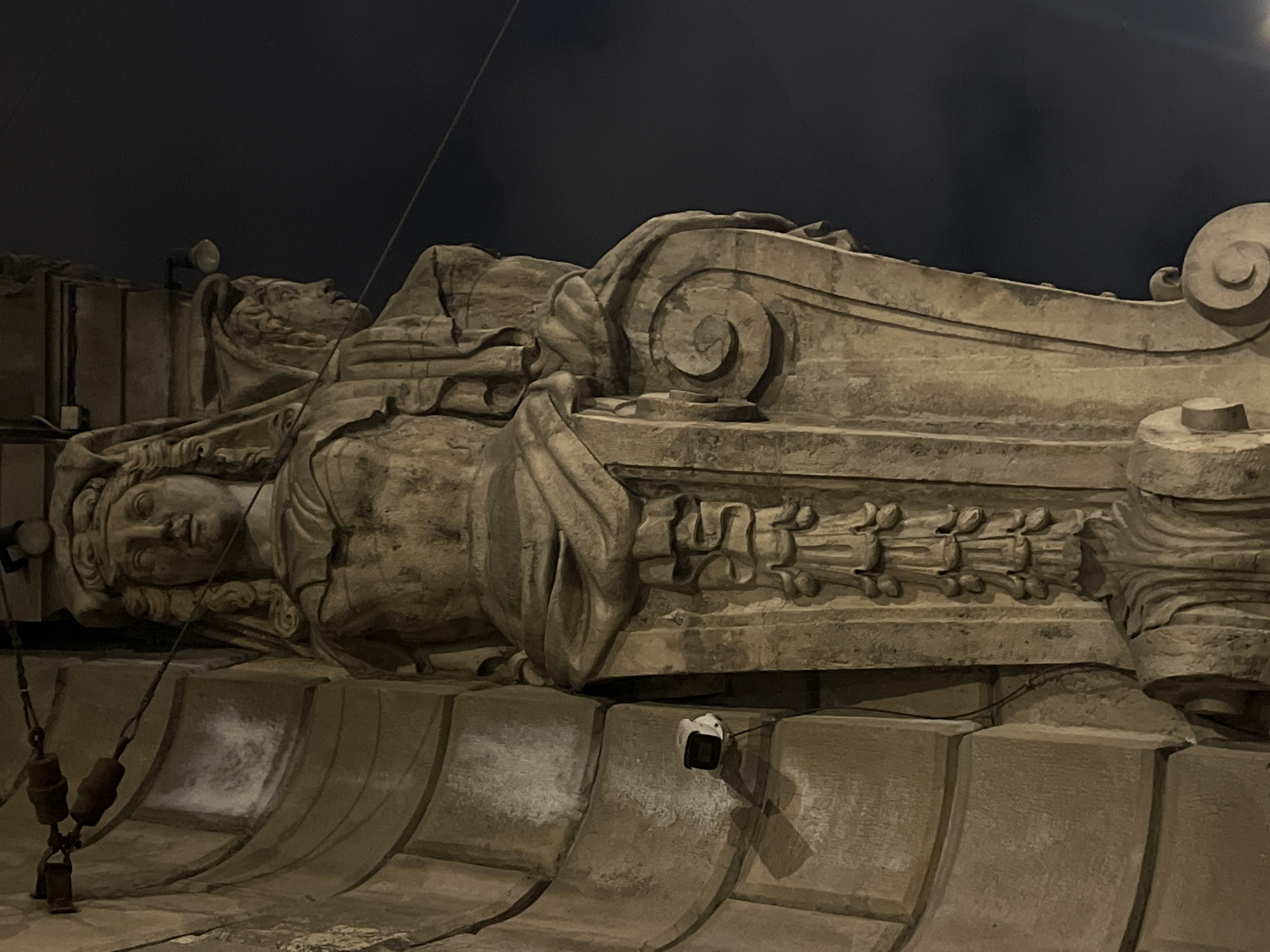 The image shows a stone sculpture of a reclining figure, possibly female, adorned with drapery. The sculpture is positioned atop an architectural structure with repeating curved elements and decorative carvings. A security camera is mounted on this structure. The scene is dimly lit, suggesting nighttime or low-light conditions. Based on the architectural style and elements, the location is likely within the city of Saint Petersburg, Russia, known for its classical architecture and sculptures.