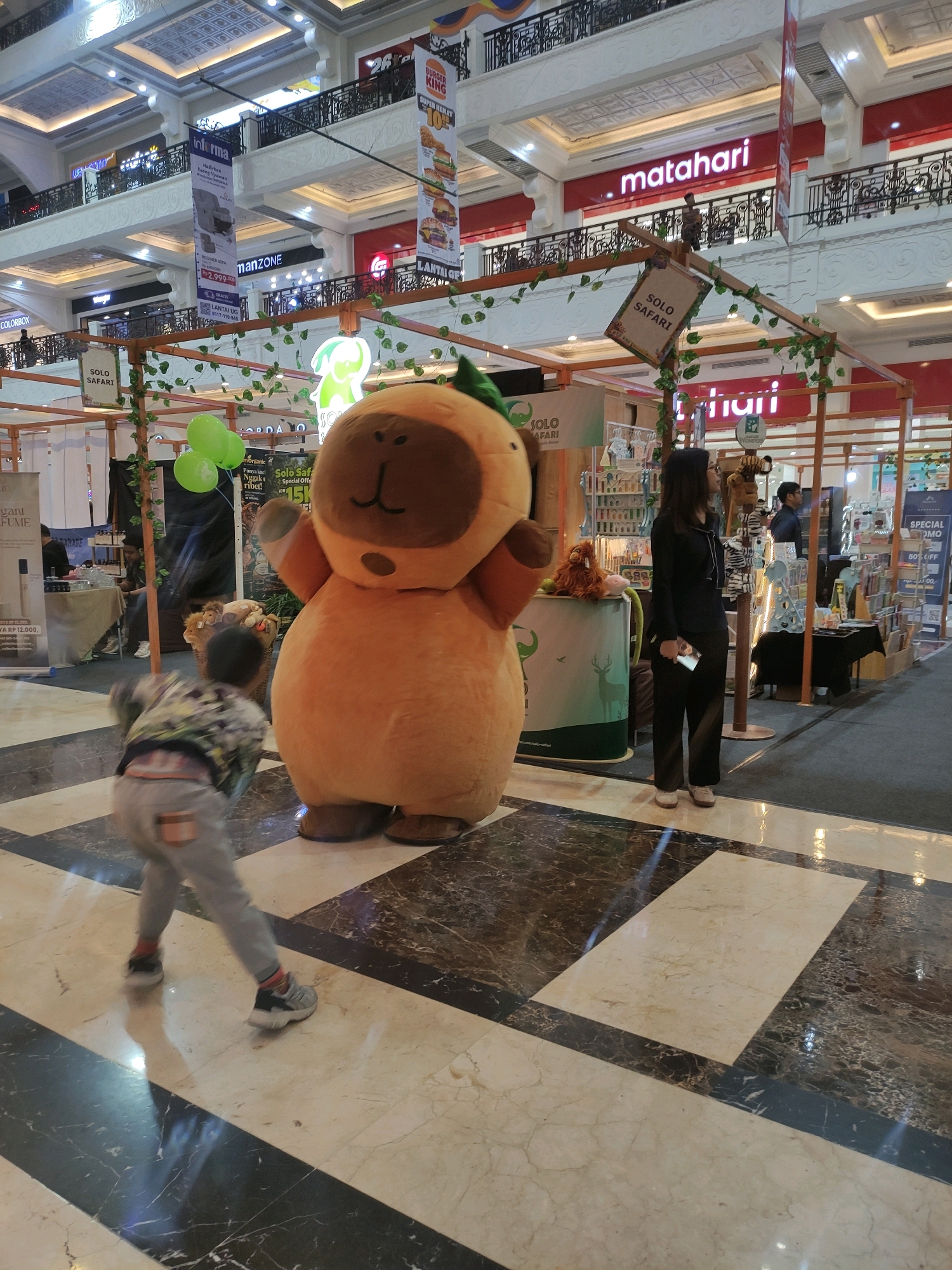 A wide-angle, eye-level shot captures a vibrant indoor scene, likely within a shopping mall, in Sinduadi, Indonesia. The foreground is dominated by a large, plush orange mascot costume resembling a character with a rounded head and small ears, standing on a checkered marble floor. To the left of the mascot, a young child, blurred due to motion, is captured in mid-stride, facing away from the camera. The child wears a grey hooded jacket with a colorful pattern and grey pants.
In the midground, a woman in a black pantsuit stands facing right, holding a white smartphone. She appears to be observing something off-camera to her right. Behind her, a stall is set up with various goods displayed, including what appears to be perfumes and other retail items. A banner with the "Matahari" logo is visible on an upper level of the mall, indicating a department store. Another banner above the stall reads "SOLO SAFARI". Green balloons are attached to a wooden structure near the mascot.
The background reveals the multi-level architecture of a shopping mall, with railings, balconies, and various shop fronts. Lighting fixtures are visible on the ceiling. The overall impression is a lively commercial space with promotional activities or events taking place.