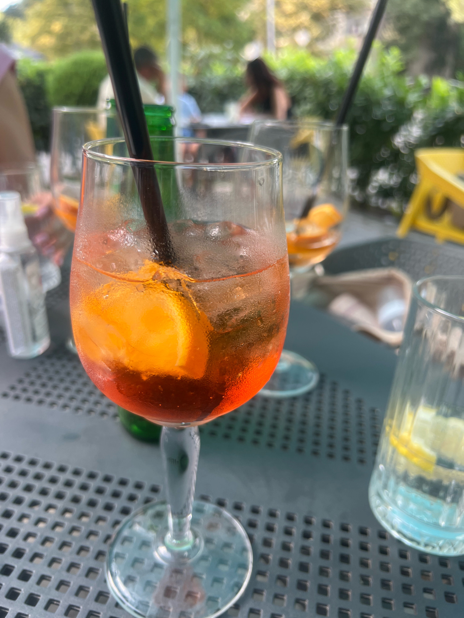 An outdoor cafe scene with an Aperol Spritz in the foreground, resting on a dark grey, perforated metal table. The drink is in a wine glass filled with ice, orange liquid, and a slice of orange, with a black straw. The glass is sweating. In the background, other drinks and glasses are visible, as well as people seated at tables, and lush green foliage. The lighting suggests daytime. The scene is consistent with a cafe setting, possibly in a city like Plovdiv, Bulgaria, known for its outdoor dining options and proximity to green spaces.