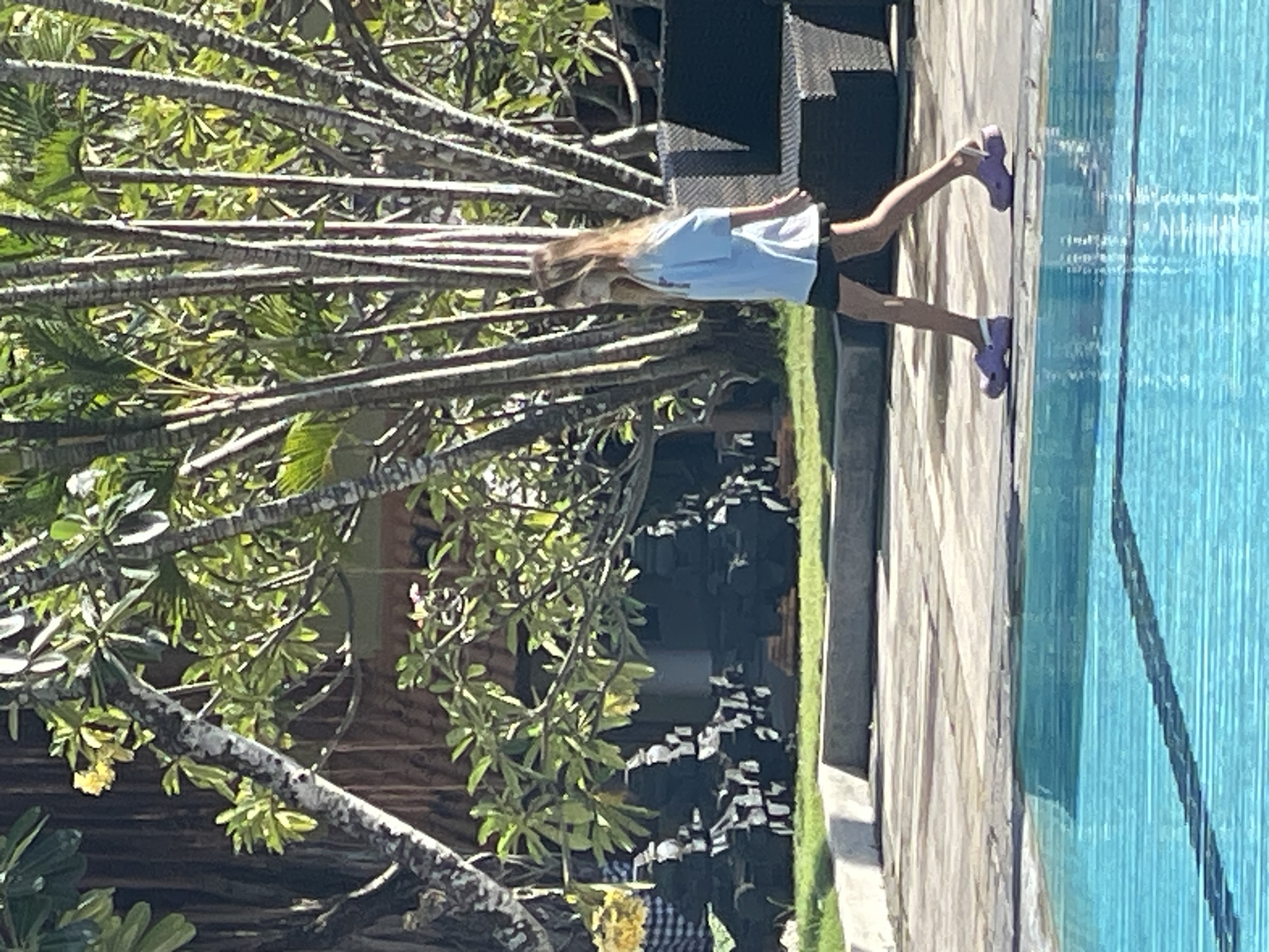 A young person with long blonde hair is walking to the left, from right to left, next to a swimming pool. They are wearing a light blue short-sleeved t-shirt and dark shorts. Their feet are clad in light purple clogs. The background features lush greenery, including palm trees and other tropical foliage, with some of the trees having distinct, textured bark. Behind the trees, a building with a thatched roof is partially visible, along with dark outdoor furniture. The ground surrounding the pool is paved with light-colored stone. The swimming pool itself is filled with bright blue water, with a dark shadow or lane line visible beneath the surface. The scene appears to be outdoors, possibly in a resort or hotel area in Kuta Selatan, Indonesia, given the tropical vegetation and pool. The lighting suggests it is daytime, with sunlight casting shadows and highlighting the foliage.