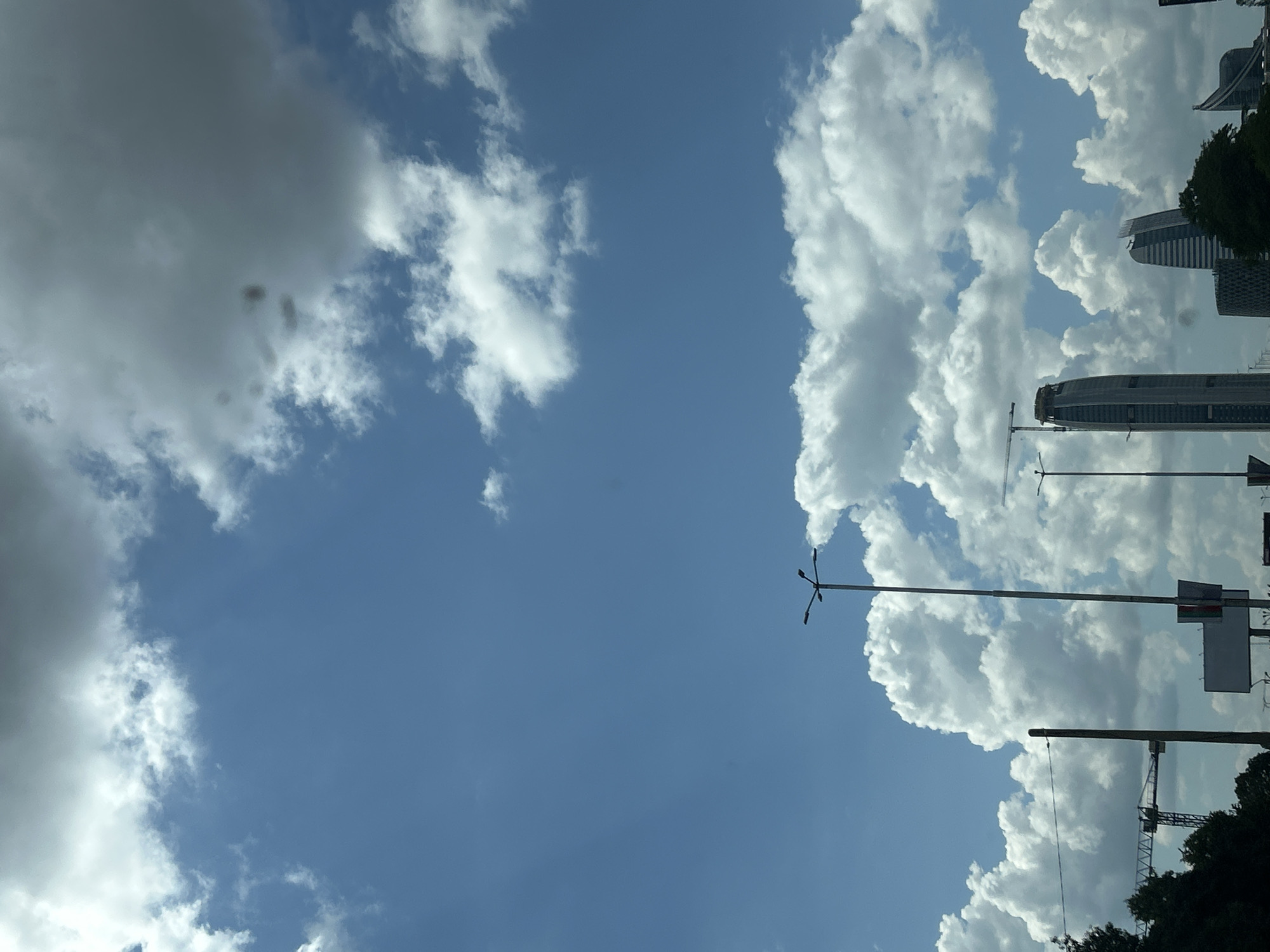 The image displays a bright daytime urban landscape under a clear blue sky dotted with numerous fluffy white cumulus clouds of varying sizes and densities; a larger, darker cloud formation is visible in the upper right quadrant. The scene is set in Sofia, Bulgaria, identifiable by the distinctive architectural structures in the lower right and center. A prominent modern skyscraper with a curved, tapering top is visible towards the center-right. Adjacent to it on the right, another modern building features a unique curved or pointed roofline, resembling a sail, and a third similar building is partially obscured further right. These structures are indicative of the Capital Fort and Sky Fort complex. Several tall streetlights with multiple fixtures extend vertically into the frame, with one prominent streetlight positioned centrally in the lower half. On the far left, a large yellow construction crane with a lattice boom is visible against the sky, indicating ongoing development. Dark green foliage and trees line the very bottom of the image on both the left and right sides. No people or vehicles are visible in the frame. The overall impression is one of a developing metropolitan area on a clear, bright day.