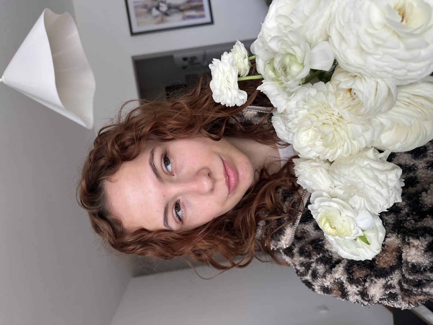 A close-up portrait captures a young woman with curly reddish-brown hair and light skin, gazing directly at the viewer with a calm, slightly smiling expression. She appears to be in her late teens or early twenties.
She is wearing a fluffy, textured jacket featuring a mottled brown, black, and cream animal print, possibly leopard or cheetah. A prominent bouquet of dense, white ranunculus flowers with numerous layered petals is positioned next to her, filling a significant portion of the right side of the frame. Some of the flowers show faint yellowish-green centers.
The background is simple, consisting of a light-colored wall, likely off-white or light grey. A white, conical or bell-shaped ceiling lamp shade hangs in the upper left corner, and a framed picture is visible on the wall further back, though its specific details are unclear. The lighting is soft, suggesting a daytime indoor setting, possibly within a home. No text is visible in the image. The scene is located in Novi Sad, Serbia.