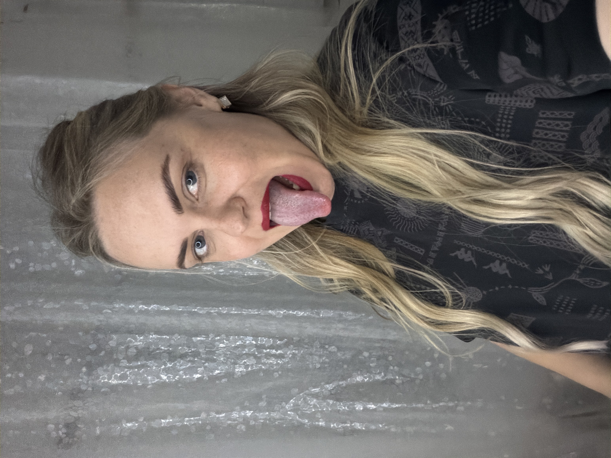 A woman with blonde hair, blue eyes, and red lipstick is sticking her tongue out. She is wearing a dark shirt with a pattern. The background appears to be a gray, textured surface. There is no information to suggest the location is Votkinsk, Russia.
