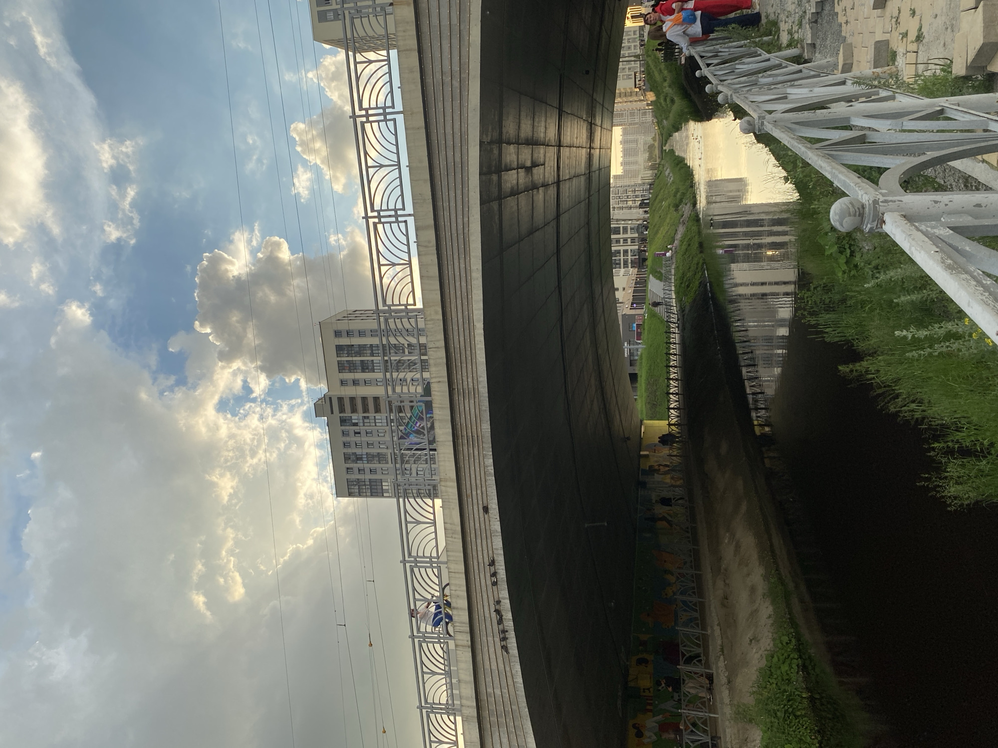 The image captures a canal flowing through a city with a bridge spanning across it. The canal is dark and reflects the sky and surrounding buildings. On the left bank of the canal, there is a grassy area with a path and a mural painted on the underside of the bridge. Several people are visible on this path, some sitting and others walking. On the right bank, there is a paved area with a white railing and more people standing, some of whom appear to be tourists.
The bridge itself is constructed of concrete and has a metal railing with decorative arches on top. A person is seen cycling across the bridge. In the background, multiple modern, tall residential buildings are visible, suggesting an urban environment. The sky is partly cloudy with dramatic, illuminated clouds, indicating either sunrise or sunset.
Based on the visual cues and publicly available information, this scene is likely located in Yekaterinburg, Russia. Specifically, the canal with the bridge and the surrounding modern architecture are characteristic of areas developed along water channels in the city, such as the Yekaterinburg city canalization system.