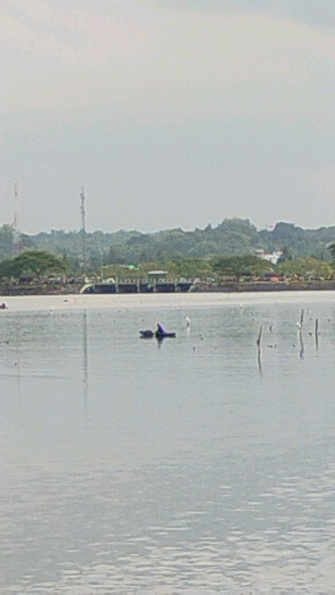 A person is on a small purple boat in the water. The person appears to be wearing a blue top and is partially submerged in the water. The boat is floating on a large body of water, possibly a lake or a bay. In the background, there is a line of trees and hills. There are also some structures that appear to be buildings and towers. The sky is overcast and appears to be a pale gray color. The scene is likely in Lhokseumawe, Indonesia, which is a coastal city in Aceh province.