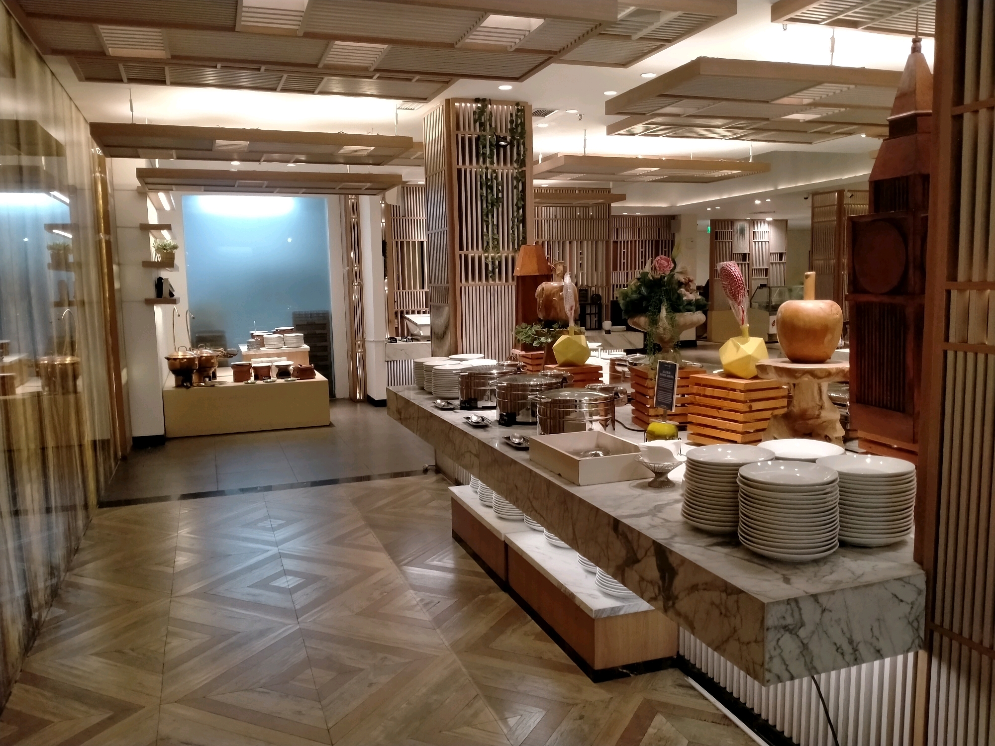 The image depicts an indoor buffet area, likely in a hotel or restaurant. The foreground features a long, marble-topped buffet counter laden with stacks of white plates, serving dishes with lids, and decorative items such as a wooden apple and a yellow geometric vase. Behind the counter, a variety of food stations are visible, with more serving dishes and decorative elements. The ceiling is adorned with a grid of wooden panels and recessed lighting. To the left, a reflective glass partition reveals a glimpse of another area with shelves and plants. The floor is covered in a light-colored wooden parquet pattern with a chevron design. In the background, wooden slatted partitions and shelves create visual separation and add to the modern, clean aesthetic. The overall atmosphere is one of a well-maintained and upscale dining establishment. While the image does not provide explicit clues to its exact location, the style is consistent with establishments found in tourist areas. If this were a restaurant in Batu, Indonesia, one might expect to see local Indonesian cuisine offerings.