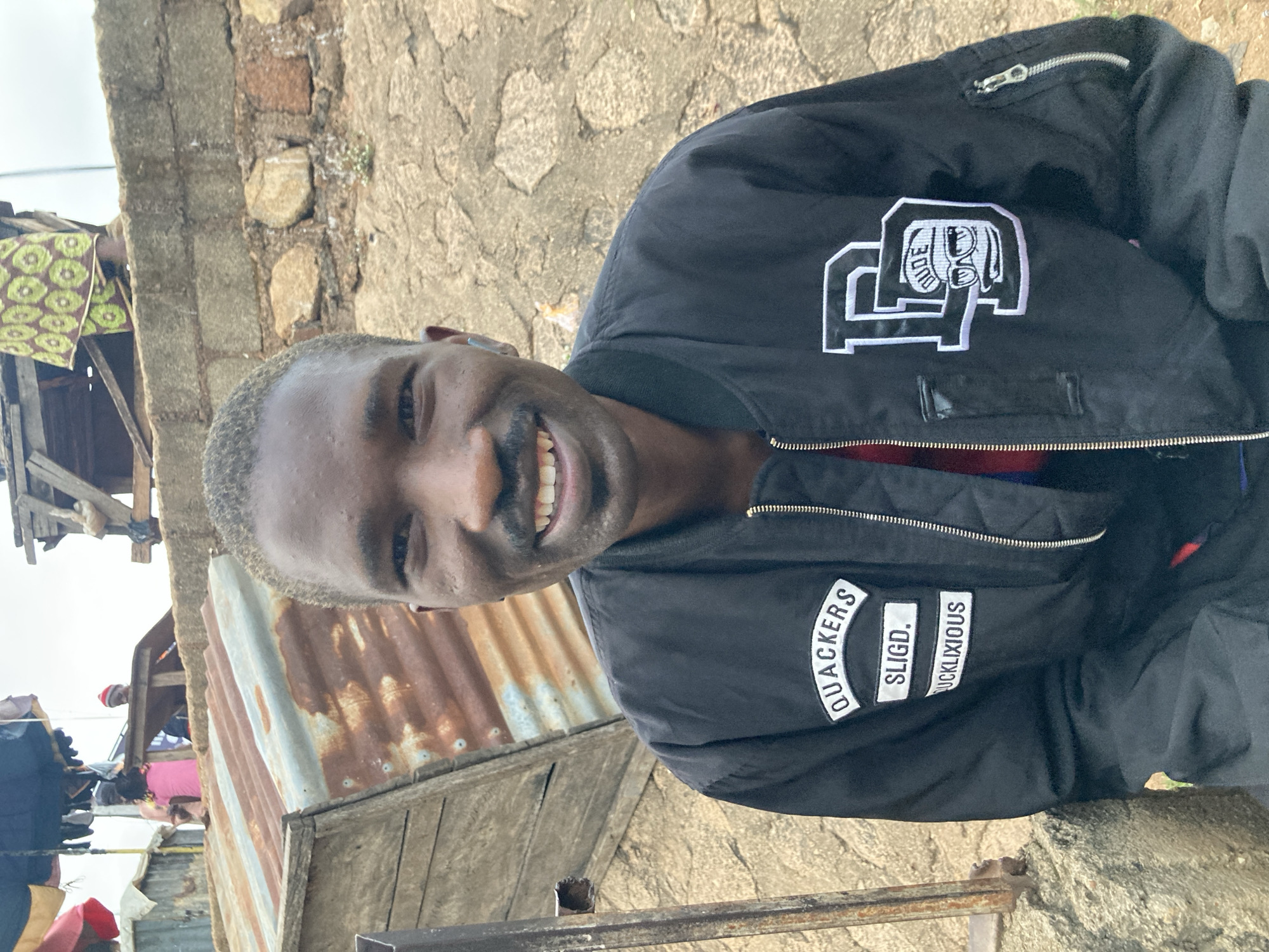 A man with short, dyed blonde hair smiles at the camera in an outdoor setting. He is wearing a black bomber jacket with white embroidered patches. One patch on the left chest depicts a stylized drawing of a figure with the word "DUDE" above it. Another patch on the right side of the jacket reads "QUACKERS SLIGD. UCKLIXIOUS". The background consists of a rough stone wall, corrugated metal sheeting, and some wooden structures, suggesting a rustic or informal environment. Other people and fabric with a green and white geometric pattern are visible in the background, hinting at a community gathering or market. The lighting suggests it is daytime, possibly under an overcast sky. The man's expression conveys happiness and friendliness. The location context indicates Gwong, Nigeria.