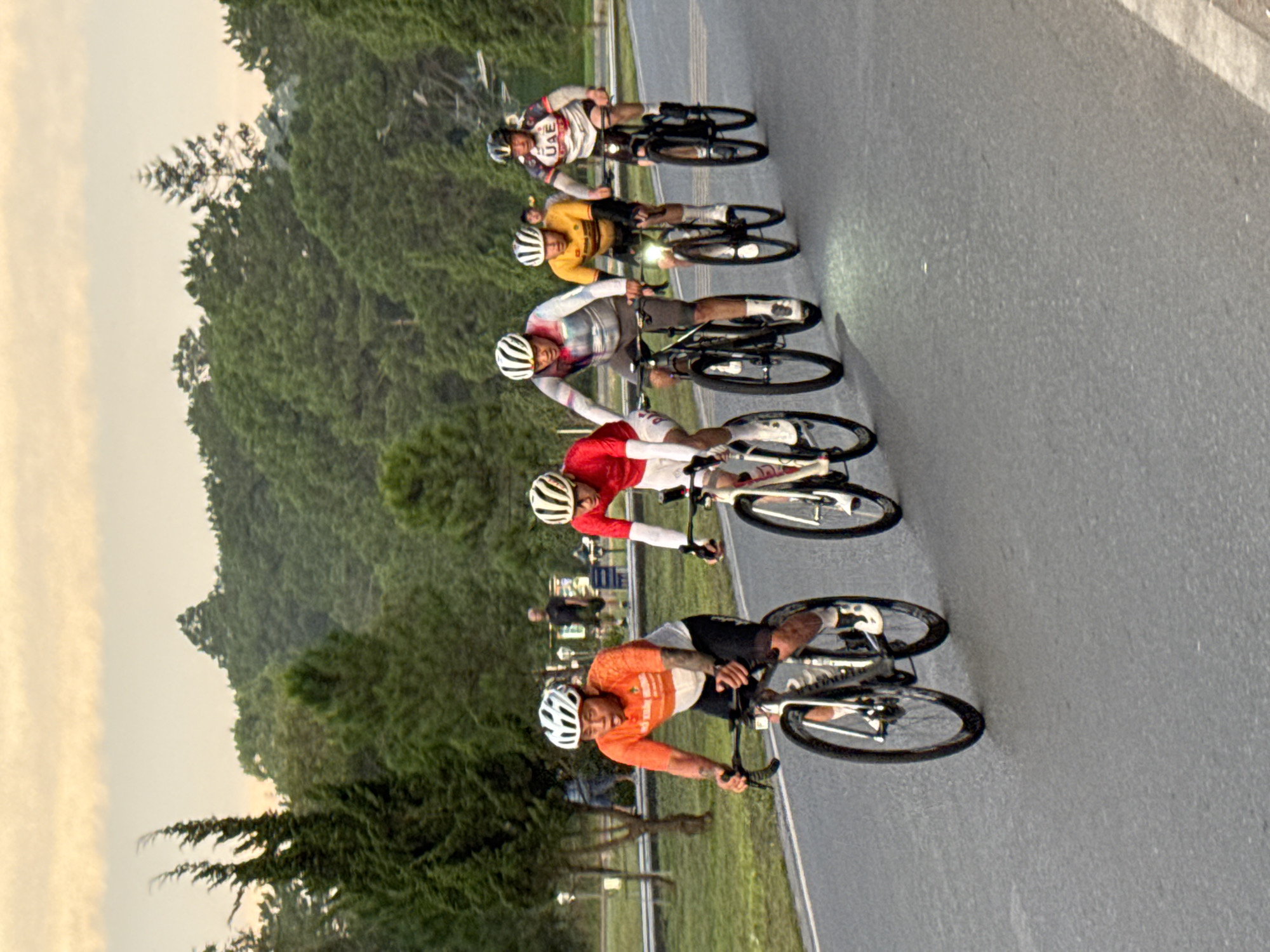 In Phường Xuân Hương - Đà Lạt, Vietnam, five cyclists, wearing helmets and racing attire, are engaged in a road cycling race. The cyclist in the foreground on the left wears an orange and white jersey and black shorts. To his right, another cyclist in a red jersey and white shorts is visible. Behind them, three more cyclists are in pursuit. The lead cyclist in the center wears a multi-colored patterned jersey, the cyclist to his right wears a yellow and black jersey, and the cyclist on the far right wears a white jersey with "UAE" emblazoned on it. They are riding on an asphalt road that curves to the right. The background features a dense forest of pine trees, with a pale, overcast sky above. The lighting suggests it is late afternoon or early morning.