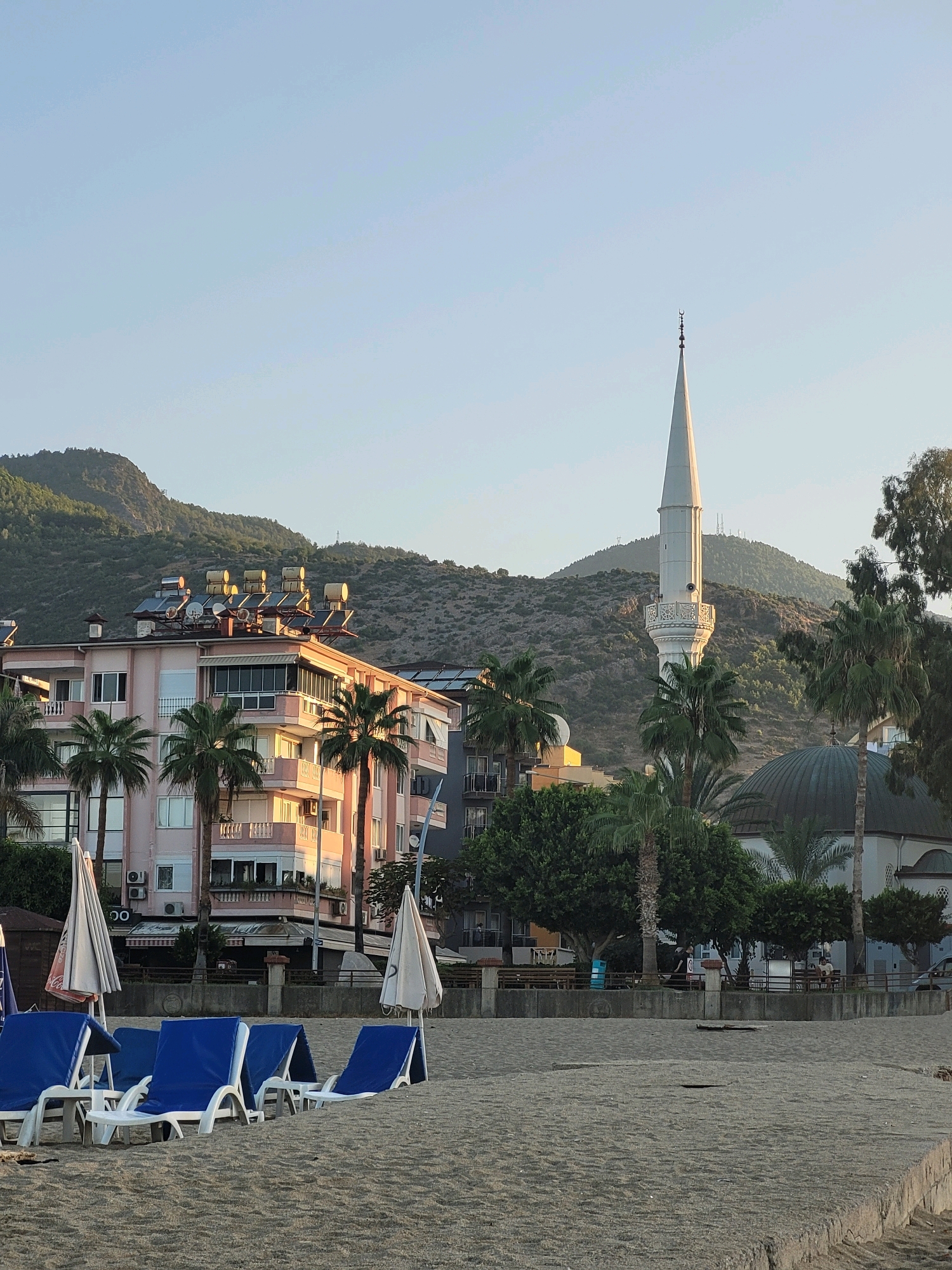 The image depicts a tranquil beach scene in Alanya, Turkey, under a clear, light blue sky, likely during the early morning or late afternoon given the soft lighting. In the foreground, several blue and white lounge chairs are neatly arranged on a sandy beach. Some are partially occupied by white beach umbrellas, suggesting preparation for a day at the beach.
Behind the loungers, a concrete promenade runs along the beachfront. Beyond the promenade, a row of buildings is visible. The most prominent is a pastel pink building with multiple balconies and solar water heaters installed on its roof. This building appears to be a residential or hotel complex. To the right of this building, a minaret of a mosque rises high into the sky, topped with a crescent moon symbol. Adjacent to the minaret is the green dome of the mosque. Palm trees are scattered throughout the scene, adding to the tropical atmosphere.
In the background, a lush, green, and mountainous landscape forms a dramatic backdrop to the coastal town. The slopes are covered with vegetation and some structures are visible on the hills, possibly communication towers. The overall impression is one of a peaceful resort town, with a blend of urban development and natural beauty.