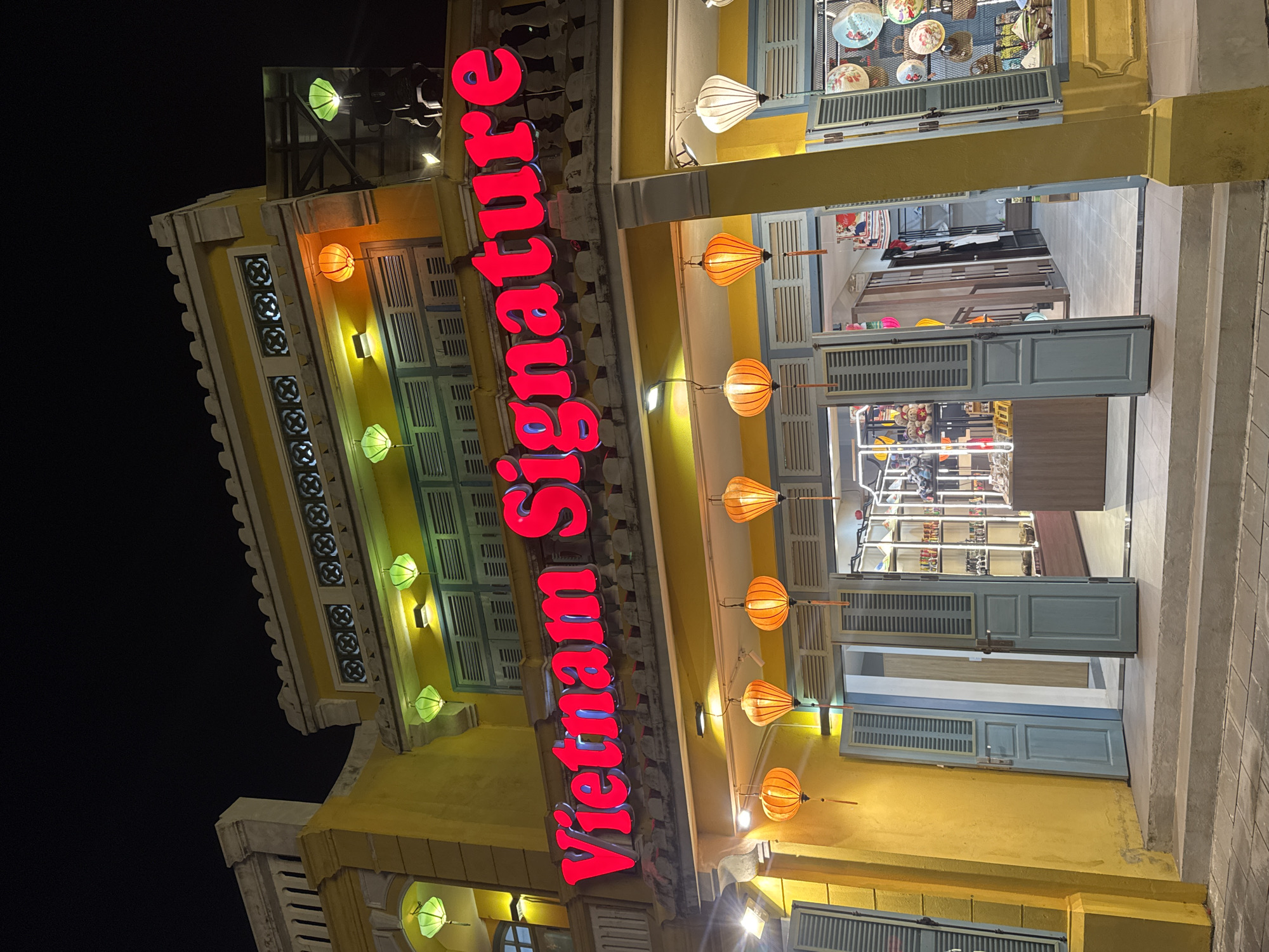 The image depicts a street view in Hội An Ward, Vietnam, showcasing a yellow building with prominent red neon signage reading "Vietnam Signature." The building features traditional shutters, lanterns, and decorative elements. The signage is vertically oriented and illuminated against the night sky. The structure is multi-story and appears to house a business, as indicated by the shop's open doors and displayed merchandise. The environment suggests a tourist district or commercial area.