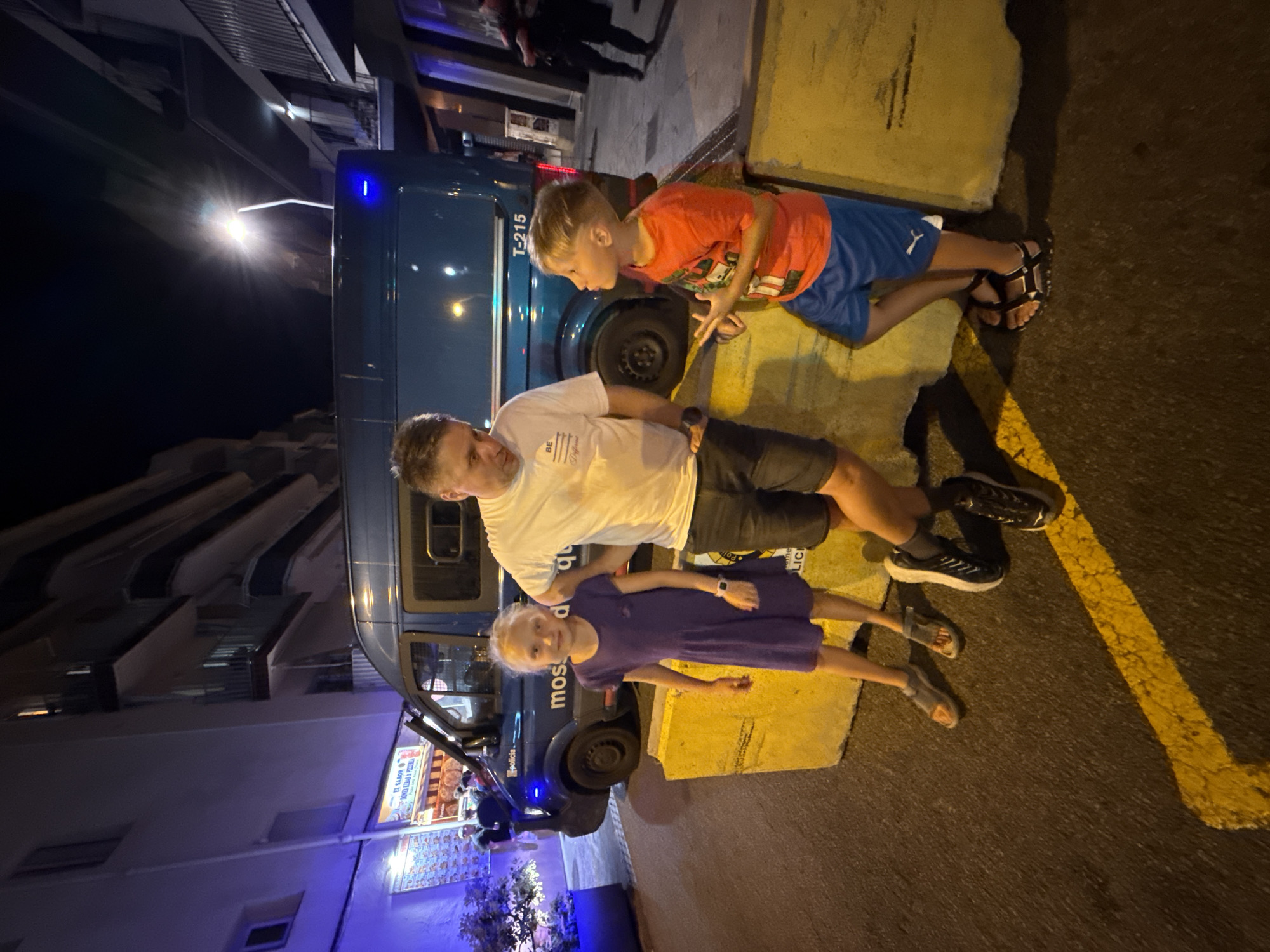 A man, a young girl, and a young boy are standing on a street at night in Blanes, Spain. The man, wearing a white t-shirt and dark shorts, is looking at the boy. The girl, wearing a purple dress, is looking at the camera. The boy, wearing an orange t-shirt and blue shorts, is looking at the man and gesturing with his hands. They are standing in front of a dark blue van with "POLICIA" written on the side. The van has a license plate that reads "T-215". The ground is asphalt, with yellow lines painted on it. There are large yellow concrete barriers behind the people. Buildings are visible in the background.