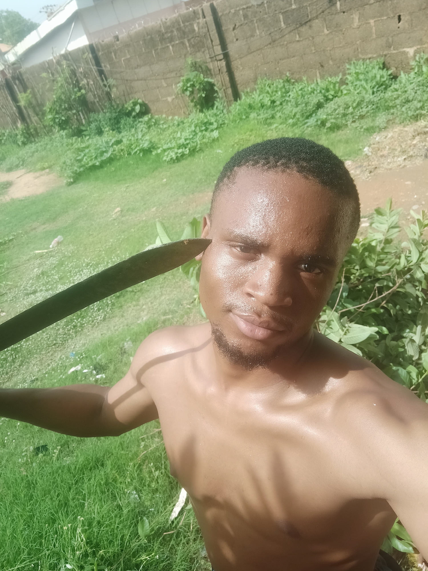 A shirtless, dark-skinned man stands in an outdoor area in Narayi, Nigeria. He holds a machete near his head. Vegetation is in the background. A brick wall is visible behind the greenery. The man appears to be sweating.
