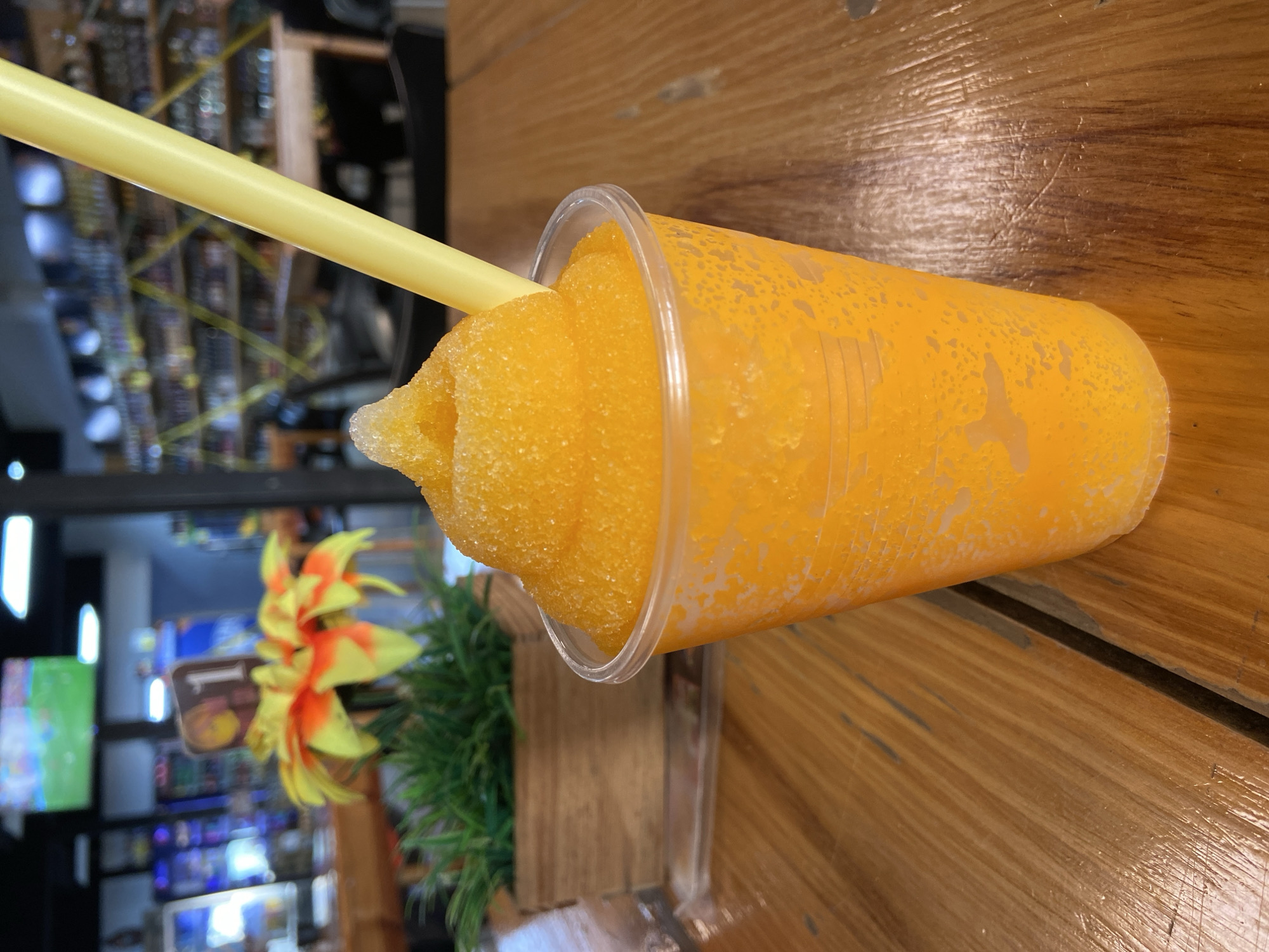 A close-up, eye-level shot shows an orange slushy in a clear plastic cup with a yellow straw, positioned on a wooden table. The slushy is piled high, forming a peak with a small amount of ice on top. The cup's exterior has condensation. In the blurred background, a wooden planter with artificial green grass and an orange and yellow flower is visible. Further back, a table with a number "1" sign is seen. The overall setting appears to be an establishment in Juan Griego, Venezuela, possibly a restaurant or bar, with shelves stocked with bottles visible in the distant background, and a television screen displaying a sports game. The lighting is artificial, and the environment is dimly lit.