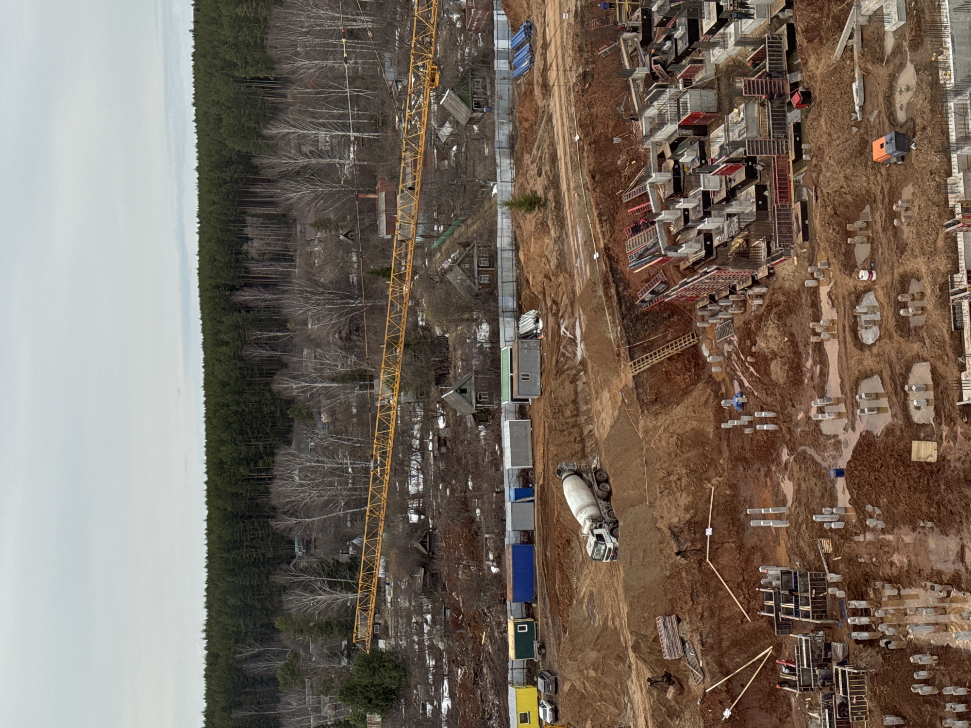 The image depicts an active construction site located in Izhevsk, Russia. A dense forest borders the left side of the frame, juxtaposed with the construction activity on the right. A yellow crane stands near a row of colorful container-like structures, likely used for storage or site offices. A concrete mixer truck is positioned near the containers. The construction site reveals foundations being laid, with numerous concrete structures and formwork visible. The earth is muddy, indicating recent excavation or damp conditions. The overall scene showcases a large-scale development in progress.