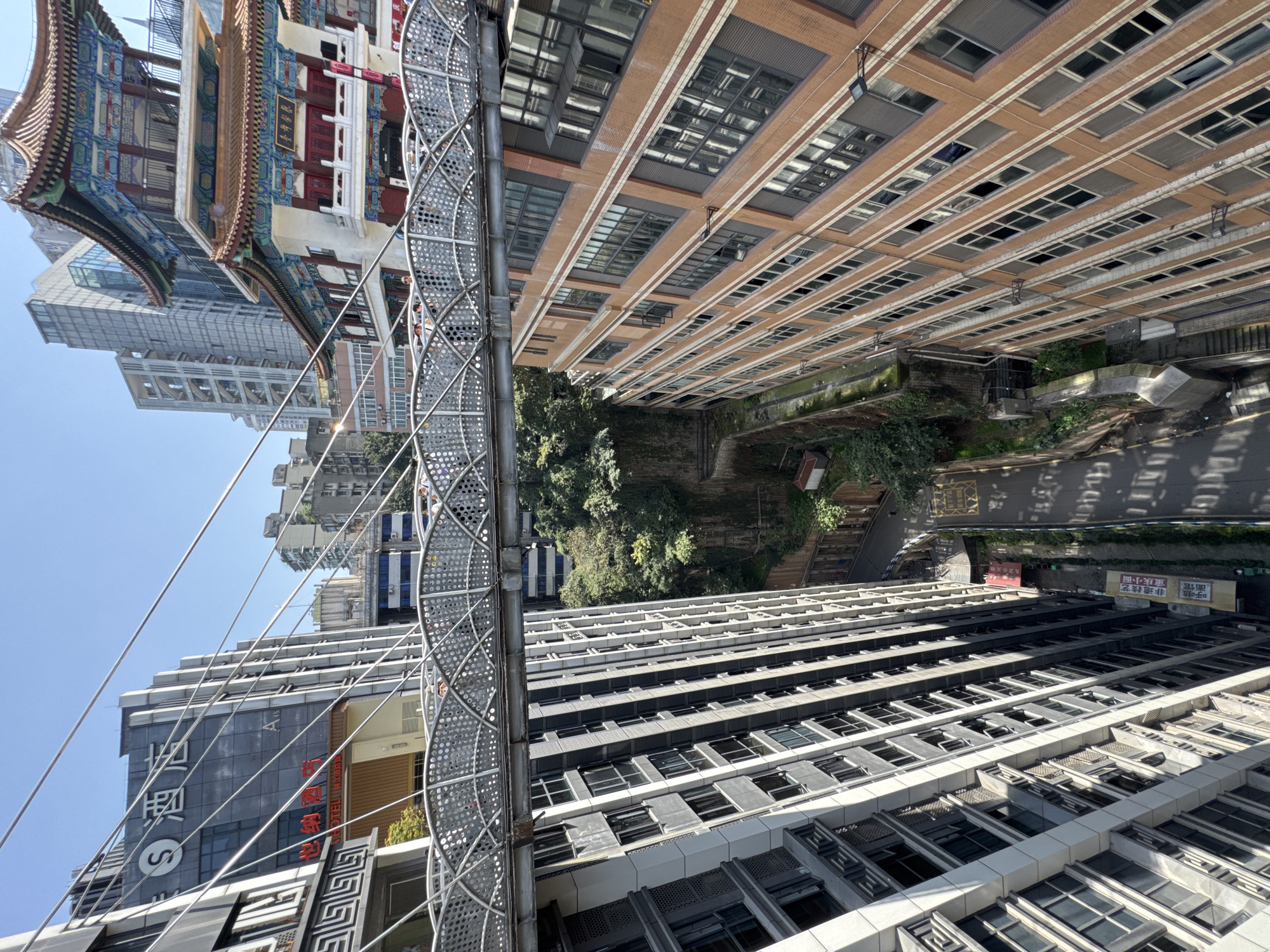 The image shows an upward-angled view of a densely populated urban area, likely in Yuzhong District, China. A modern metal pedestrian bridge bisects the image diagonally, connecting buildings. On the left side of the bridge is traditional Chinese architecture with ornate detailing and a multi-tiered roof. Modern high-rise buildings are also visible in the background. On the right side of the bridge, there is a brick building with rows of windows, along with a narrow street and some greenery. The overall scene depicts a blend of traditional and modern architecture within a bustling city environment.